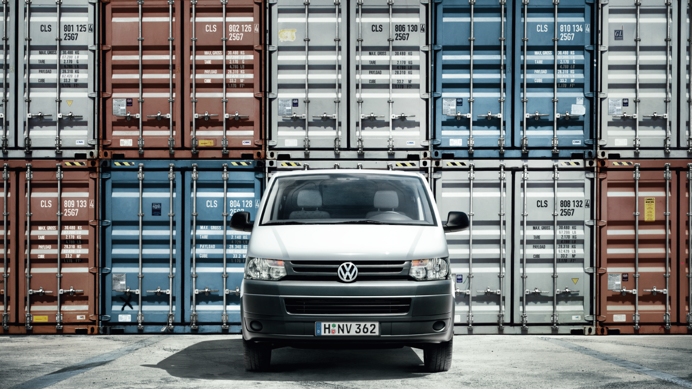 Biela dodávka značky Volkswagen stojí pred množstvom farebných lodných kontajnerov na nákladnú dopravu. Na evidenčnom čísle vozidla je kód „H-NV 362“. Povrch, na ktorom vozidlo parkuje, je asfaltový.