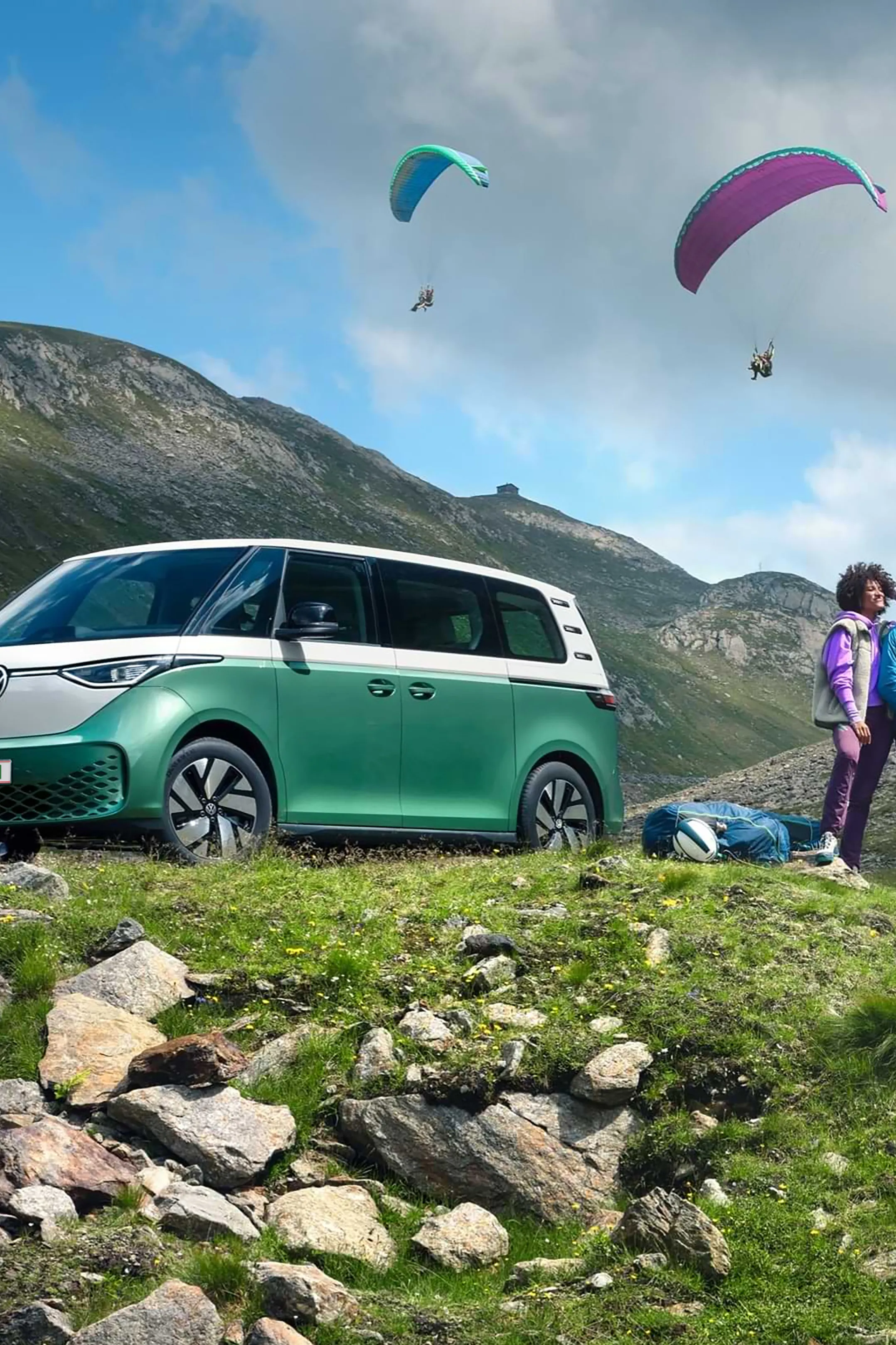 Dvojica stojí na zelenej lúke vedľa zeleného VW ID. Buzz. Na oblohe sú dvaja paraglidisti, ktorí lietajú nad kopcami. V pozadí vidieť kopce.