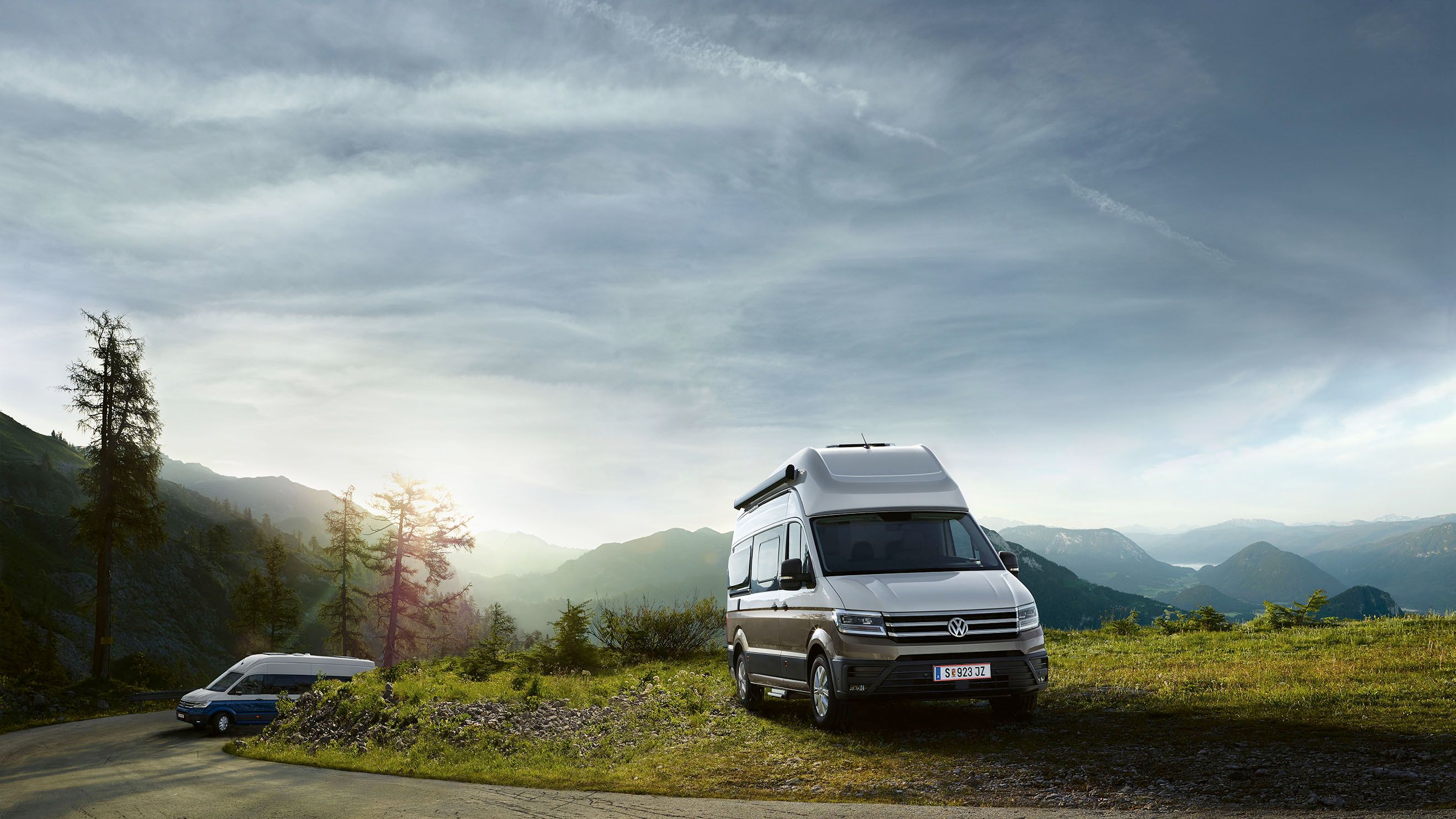 Dodávka zaparkovaná na vrchole kopca s výhľadom na hory pri východe slnka. Cesta je úzka a vedie cez zeleň. ďalšia VW California prechádza zákrutou.