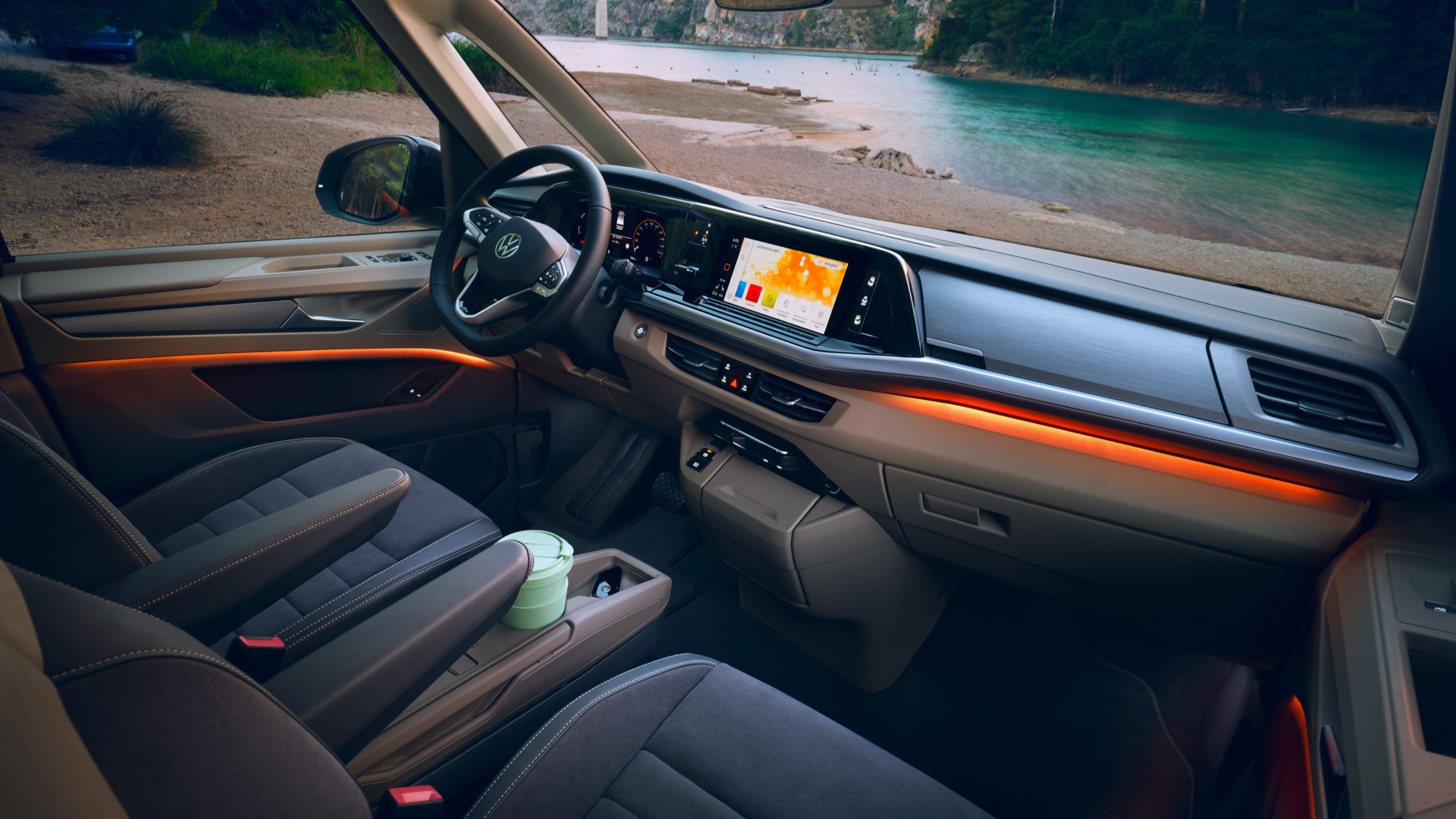 Ambientné osvetlenie VW Multivan Energetic. Pohľad do interiéru vozidla na miesto vodiča a spolujazdca, palubnú dosku a stredovú konzolu. Cez predné a bočné okno vidieť krásnu horskú rieku. Vozidlo je zaparkované na brehu rieky v horách.