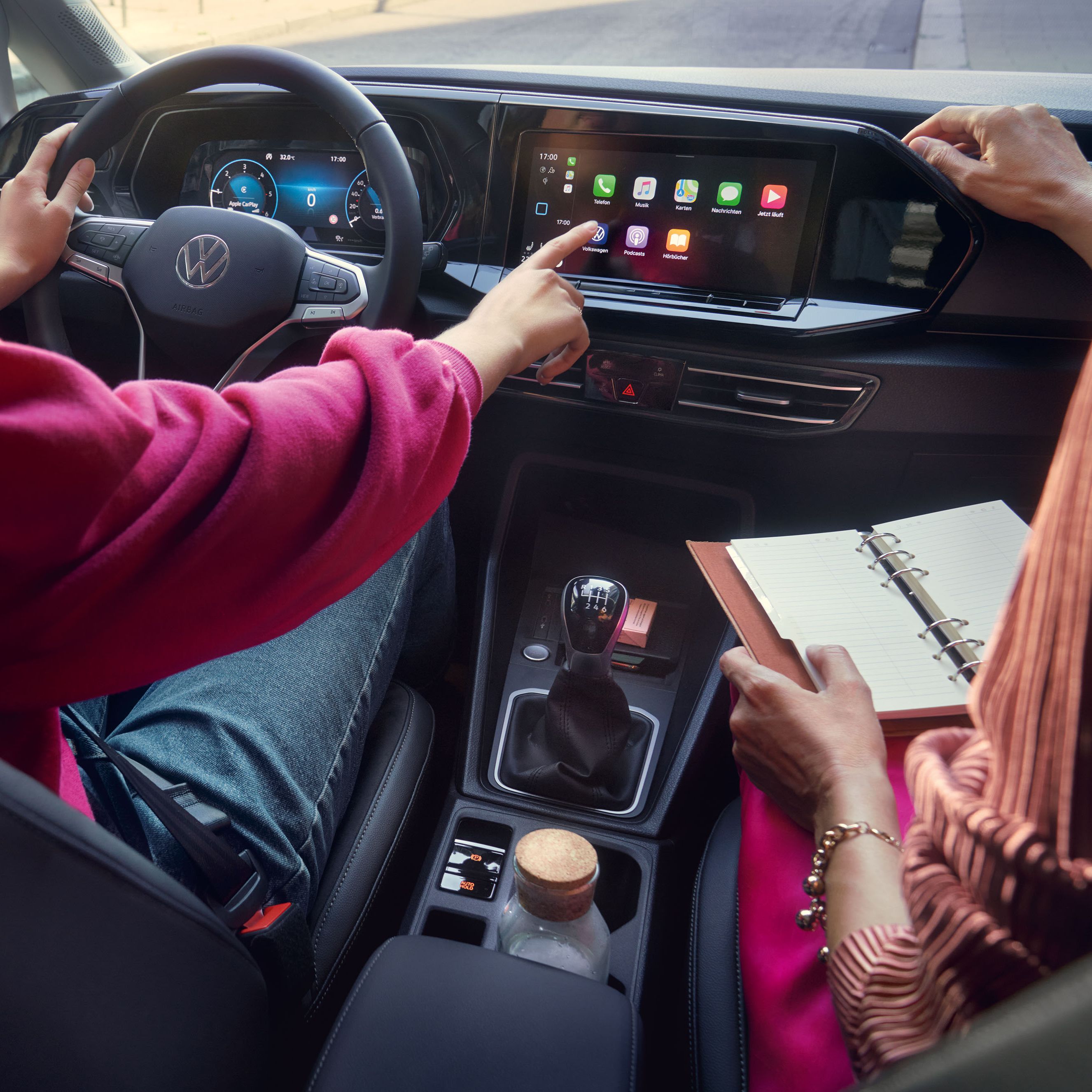 Dvaja ľudia sedia v aute značky Volkswagen. Žena na sedadle vodiča ovláda dotykový displej systému infotainment na palubnej doske. Druhá osoba drží otvorený diár na svojich kolenách.