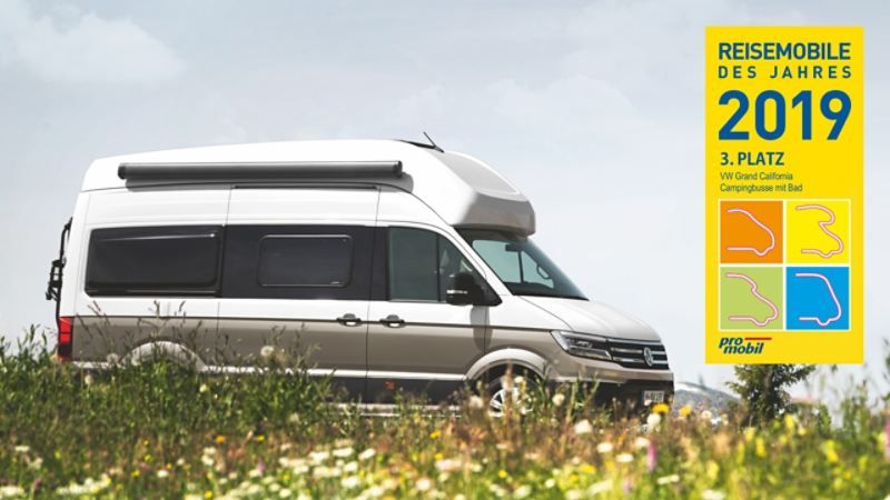 Rekreačný obytný automobil VW Grand California zaparkovaný na lúke. na obrázku je tiež grafické označenie oznamujúce tretie miesto ako "Reisemobile des Jahres 2019". Divoké kvety rastúce na lúke dodávajú obrázku prírodnú atmosféru.