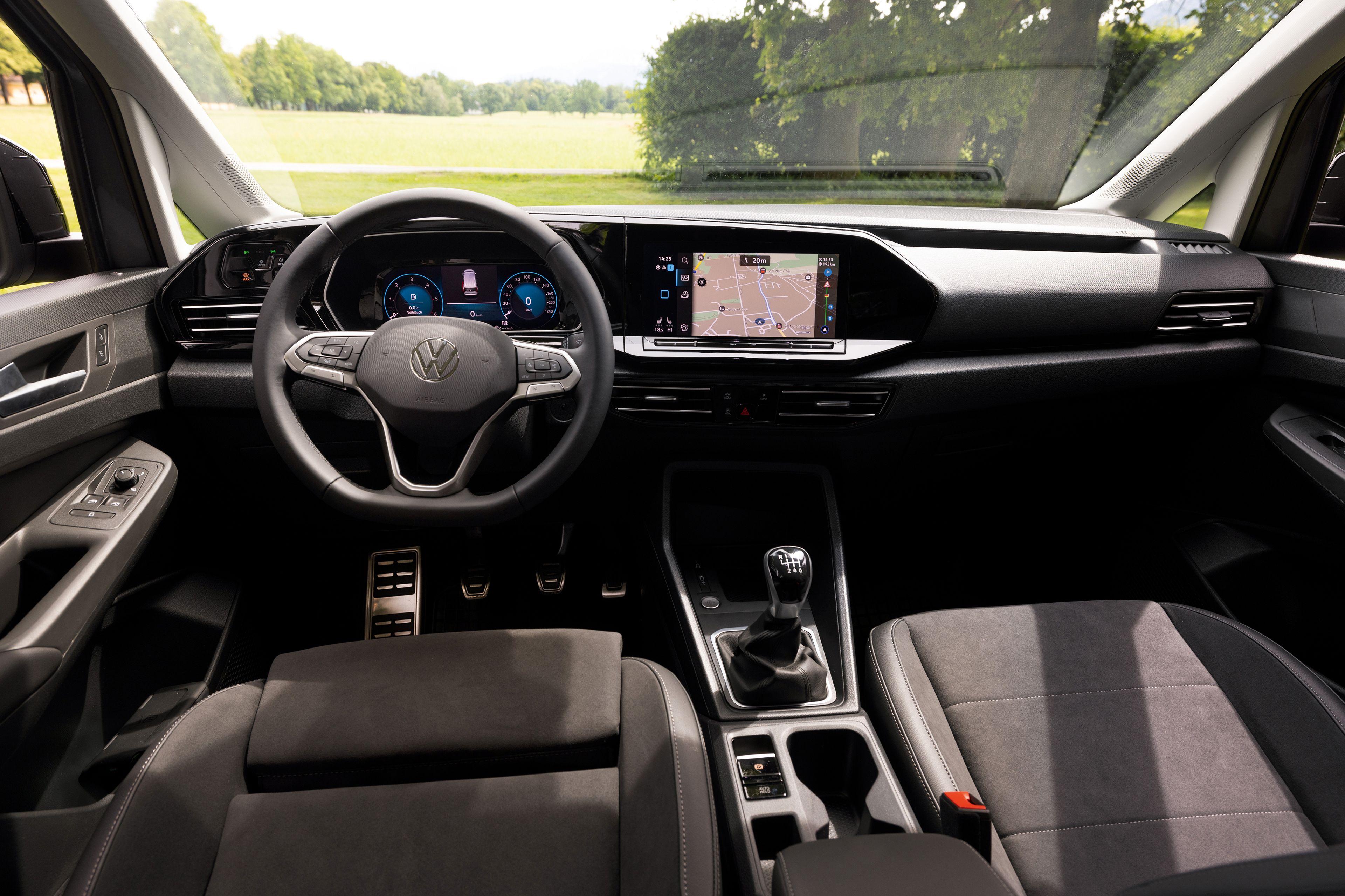 Interiér automobilu Volkswagen s logom na volante. Na palubnej doske sú dva veľké digitálne displeje, jeden prístrojového panela zobrazujúci informácie o vozidle a druhý systému infotainment s navigačným systémom. Sedadlá sú čalúnené tmavým materiálom.  Cez čelné  sklo je výhľad na rozsiahlu lúku, ktorú lemujú stromy.