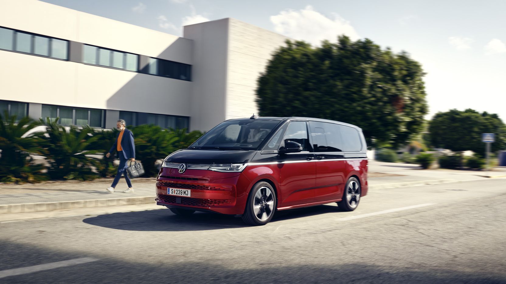 VW Multivan počas jazdy na ceste popri nízkej budove, pri kotorej je veľa zelene.