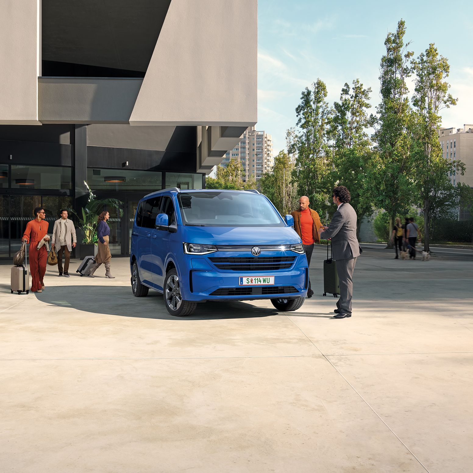 Modrý Volkswagen Caravelle je zaparkovaný pred modernou budovou so zaujímavou fasádou. Skupina ľudí s kuframi prichádza k vozidlu. Na oblohe je slnečný deň s niekoľkými oblakmi.