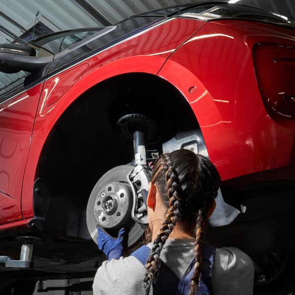 Servisná pracovníčka vykonáva údržbu na prednom kolese červeného auta, ktoré je zdvihnuté. Mechanička kontroluje brzdný kotúč a zavesenie kolies.