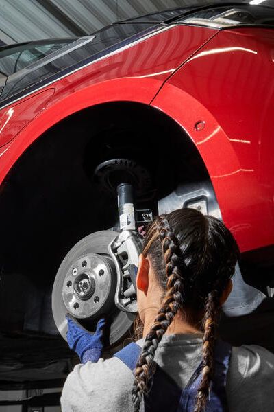 Servisná pracovníčka vykonáva údržbu na prednom kolese červeného auta, ktoré je zdvihnuté. Mechanička kontroluje brzdný kotúč a zavesenie kolies.