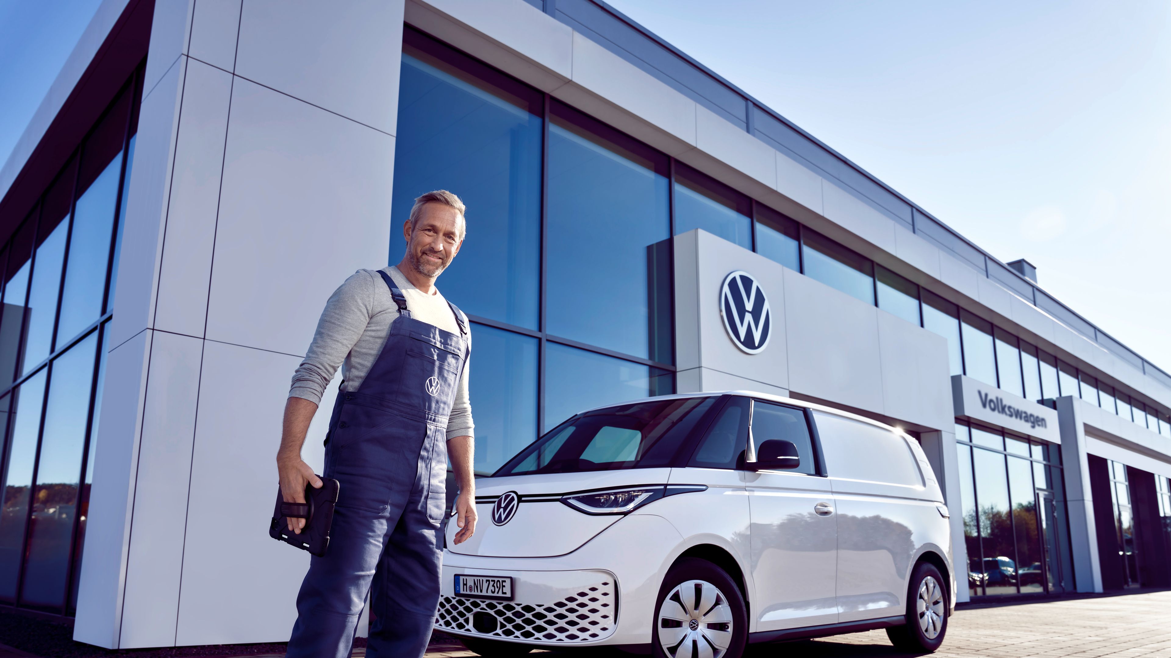 Servisný pracovník v modrých montérkach stojí pred bielym ID. Buzz, ktorí je zaparkovaný pred showroomom Volkswagen. Muž sa pozerá priamo do objektívu.