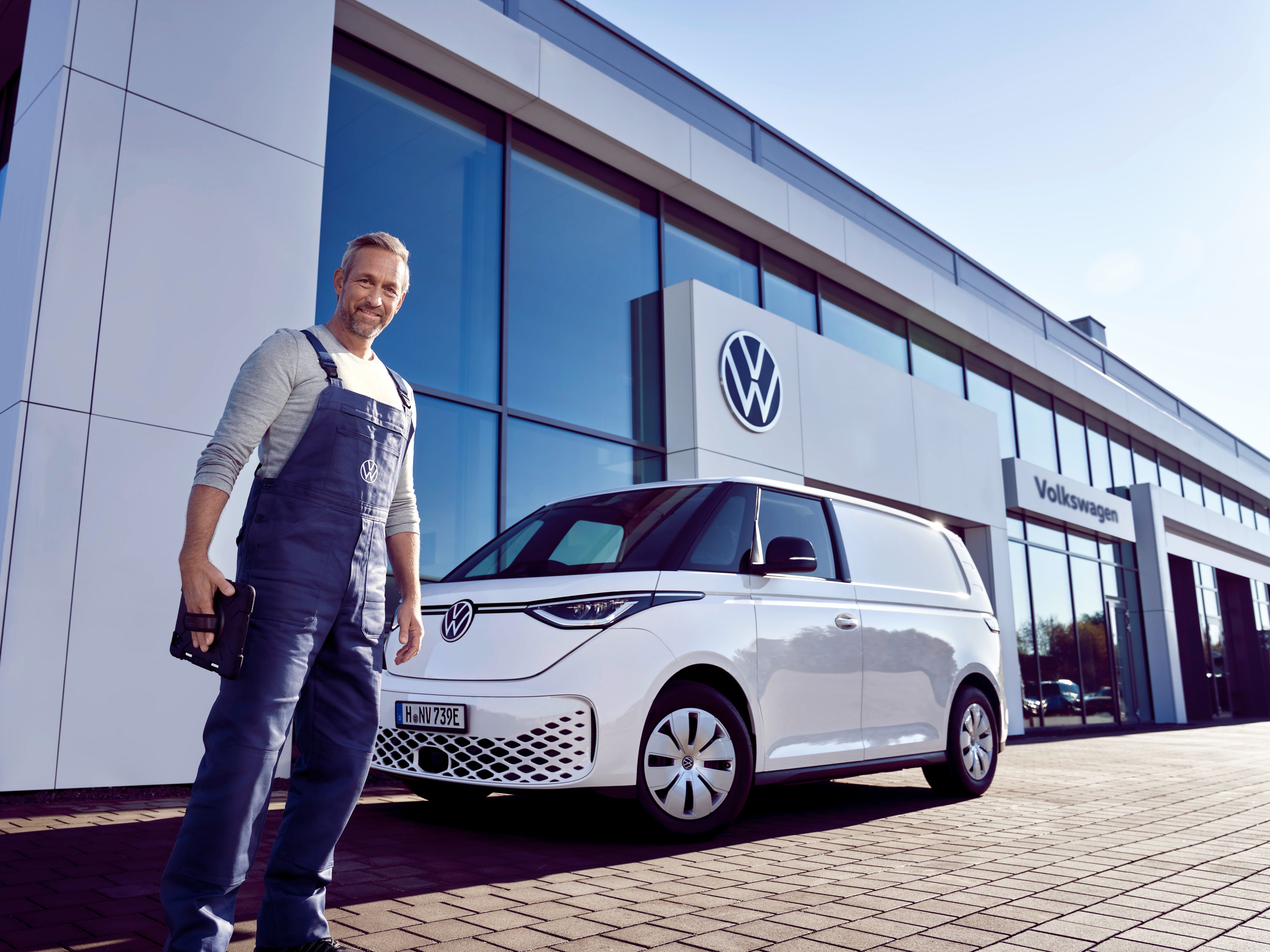 Servisný pracovník v modrých montérkach stojí pred bielym ID. Buzz, ktorí je zaparkovaný pred showroomom Volkswagen. Muž sa pozerá priamo do objektívu.