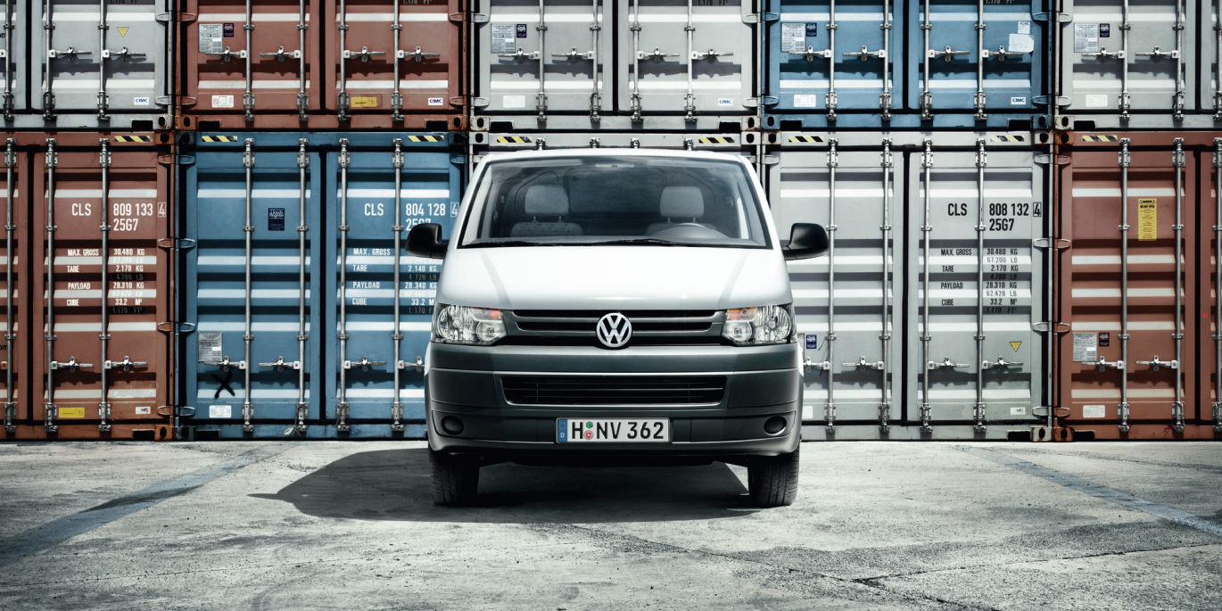 Biela dodávka značky Volkswagen stojí pred množstvom farebných lodných kontajnerov na nákladnú dopravu. Na evidenčnom čísle vozidla je kód „H-NV 362“. Povrch, na ktorom vozidlo parkuje, je asfaltový.