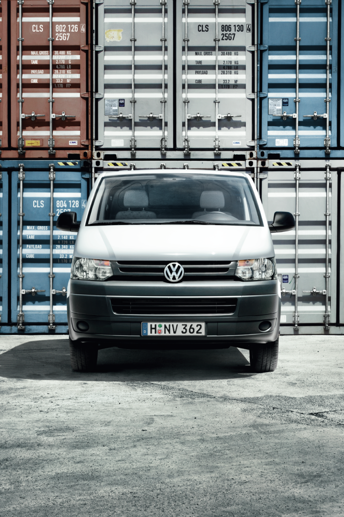 Biela dodávka značky Volkswagen stojí pred množstvom farebných lodných kontajnerov na nákladnú dopravu. Na evidenčnom čísle vozidla je kód „H-NV 362“. Povrch, na ktorom vozidlo parkuje, je asfaltový.