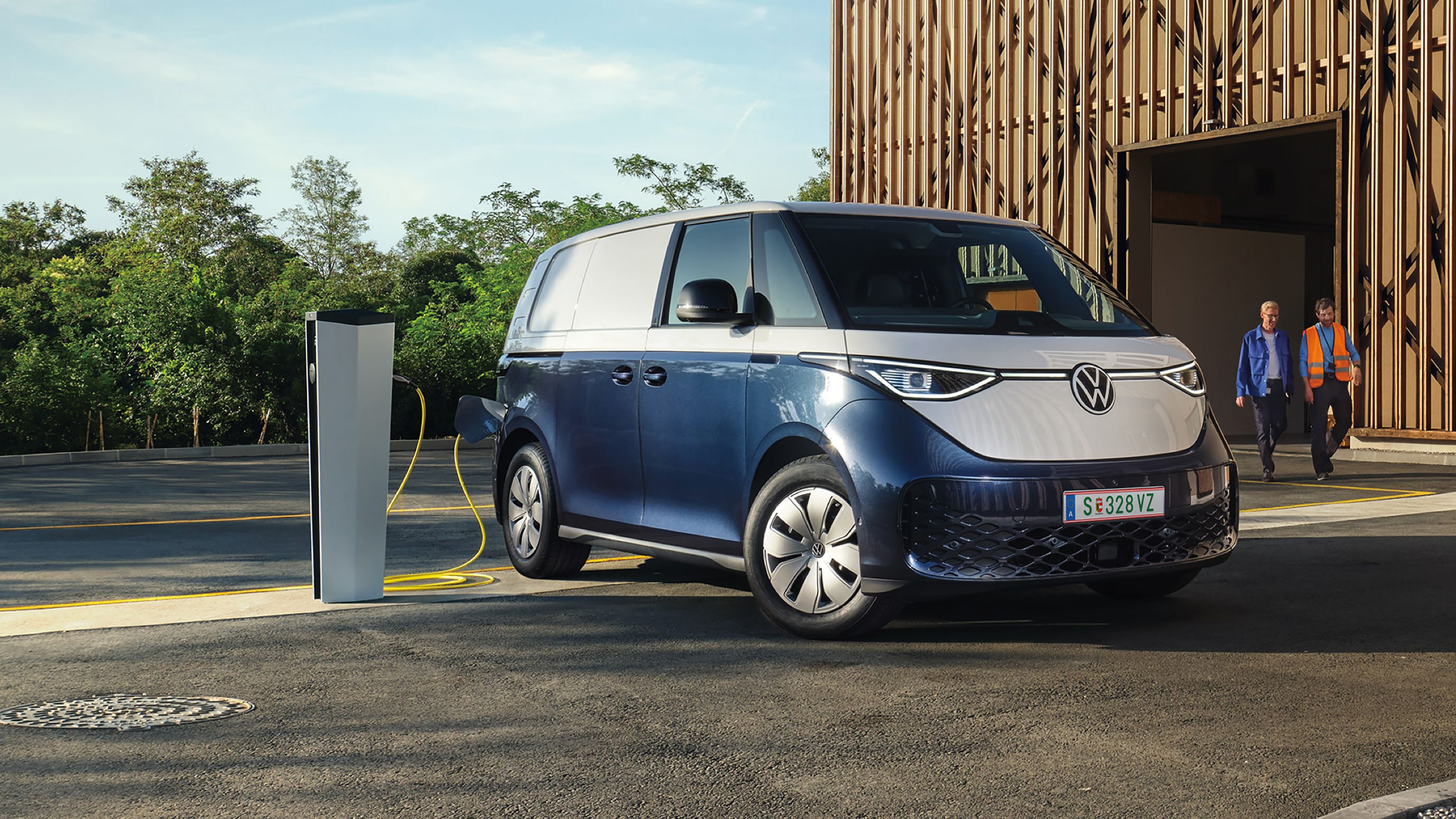Modrý elektrický Volkswagen ID.Buzz Cargo je zaparkovaný pri nabíjacej stanici a nabíja sa. V pozadí sú dvaja muži kráčajúci okolo modernej drevenej budovy. Na oblohe sú jemné mraky.