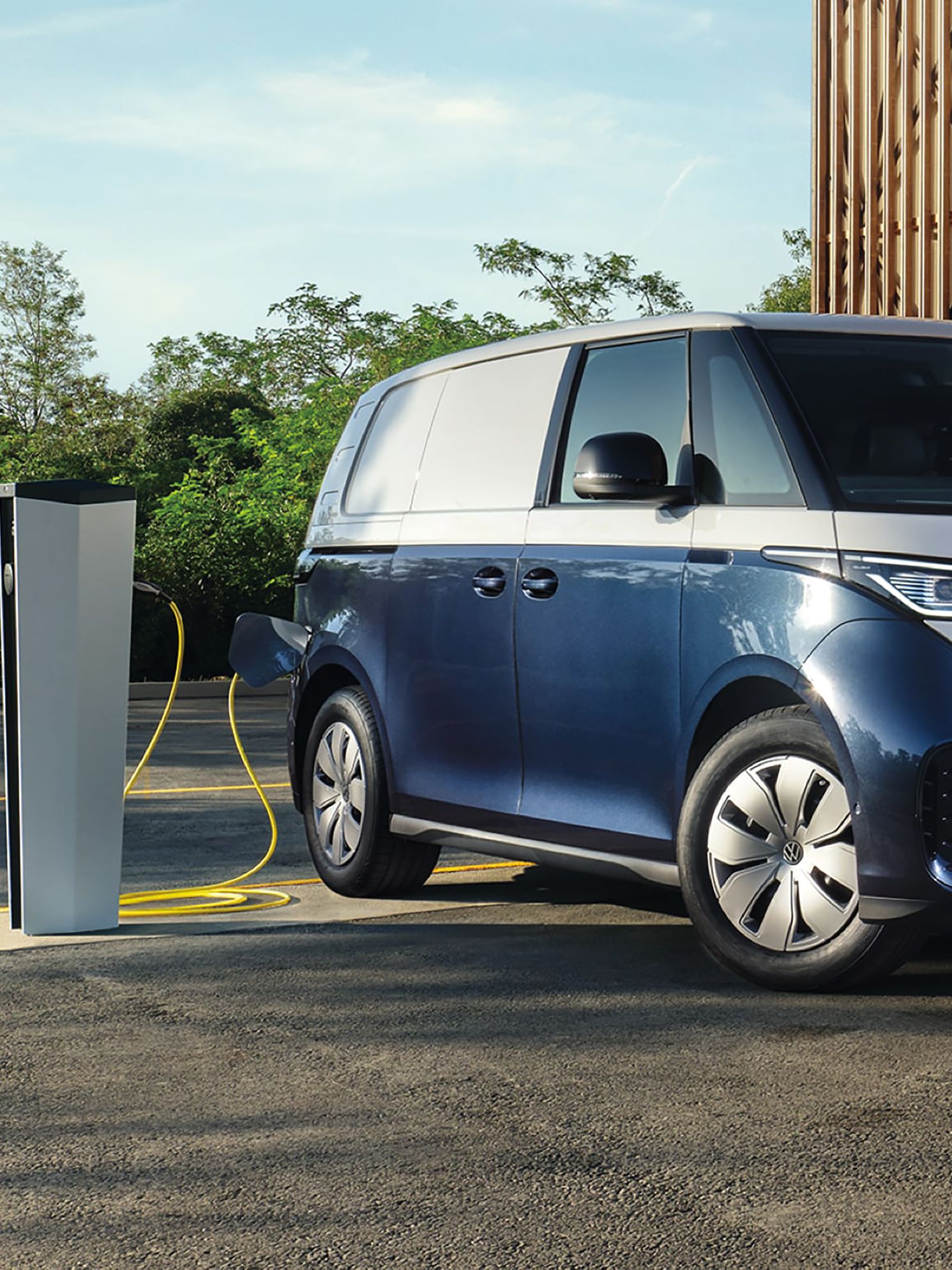 Modrý elektrický Volkswagen ID.Buzz Cargo je zaparkovaný pri nabíjacej stanici a nabíja sa. V pozadí sú dvaja muži kráčajúci okolo modernej drevenej budovy. Na oblohe sú jemné mraky.