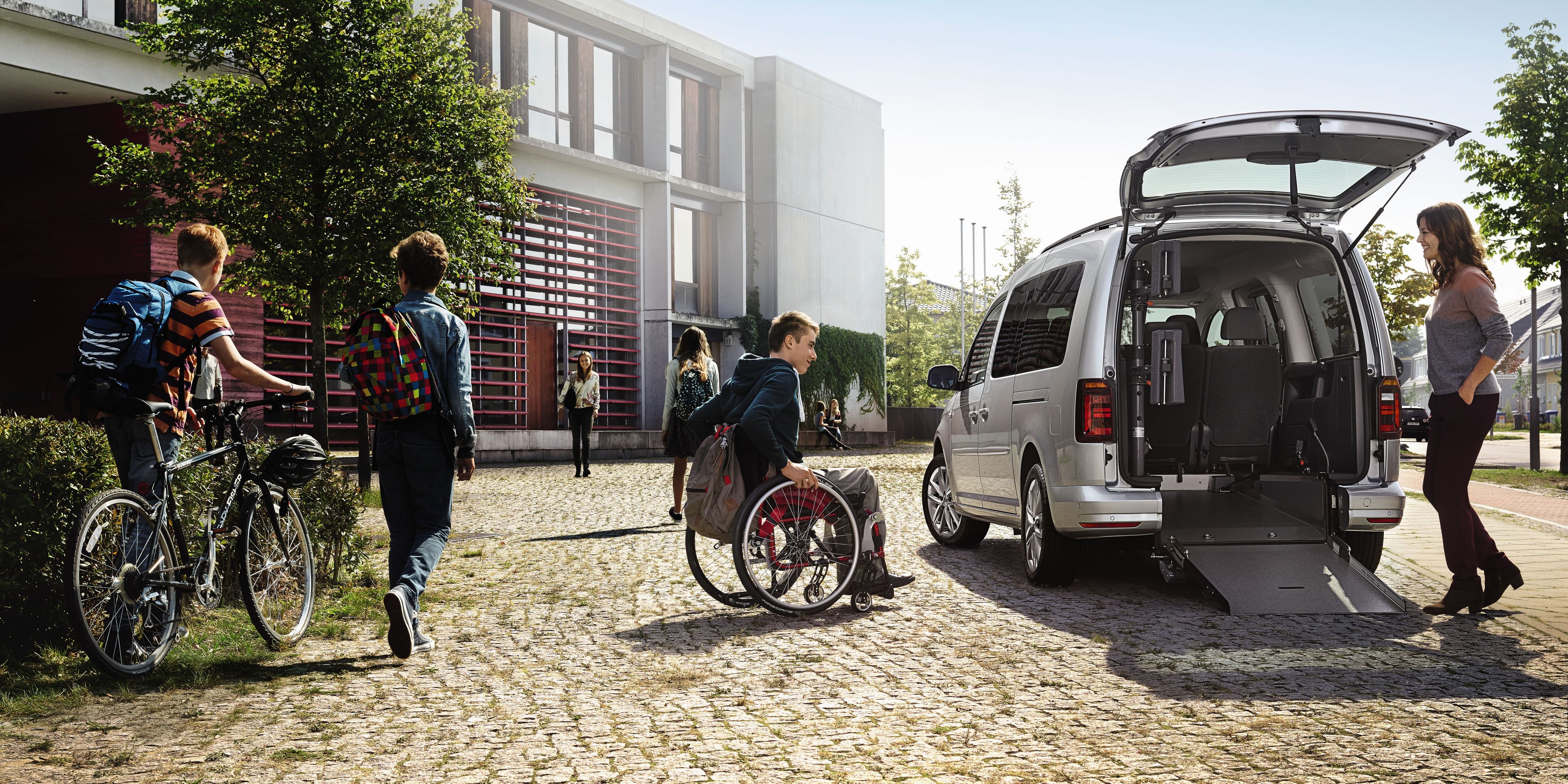 Na obrázku je niekoľko osôb, z ktorých jedna sedí na invalidnom vozíku a používa rampu na nastúpenie do auta. Zvyšné osoby vedú bicykle a blízko je aj budova obklopená stromami. 