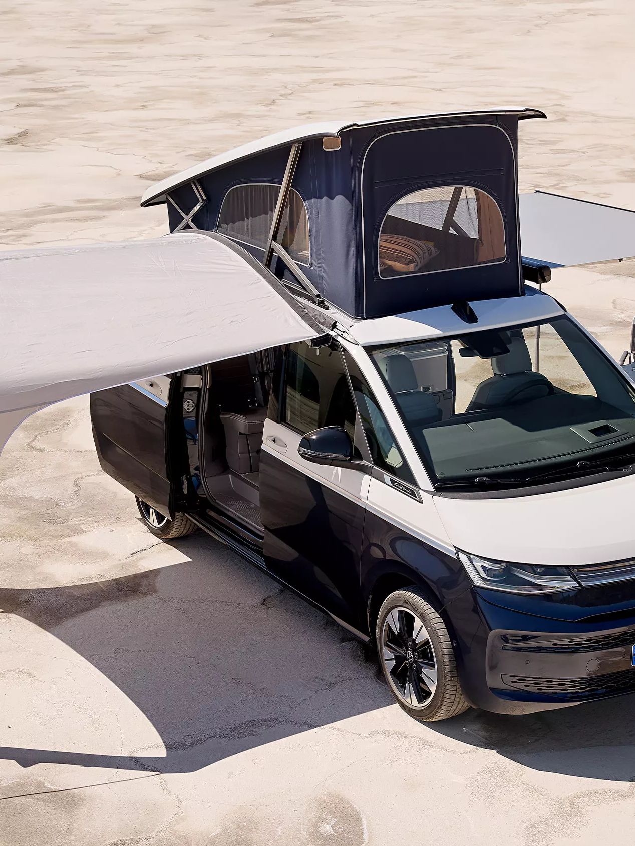Modrý obytný karavan Volkswagen California so zdvihnutou strechou stojí na rovnom povrchu. Z jedného boku vozidla je roztiahnutá markíza, pod ktorou sú dve stoličky a stôl s občerstvením. Z druhého boku je postavený prístrešok. Okolie je svetlé a pusté.