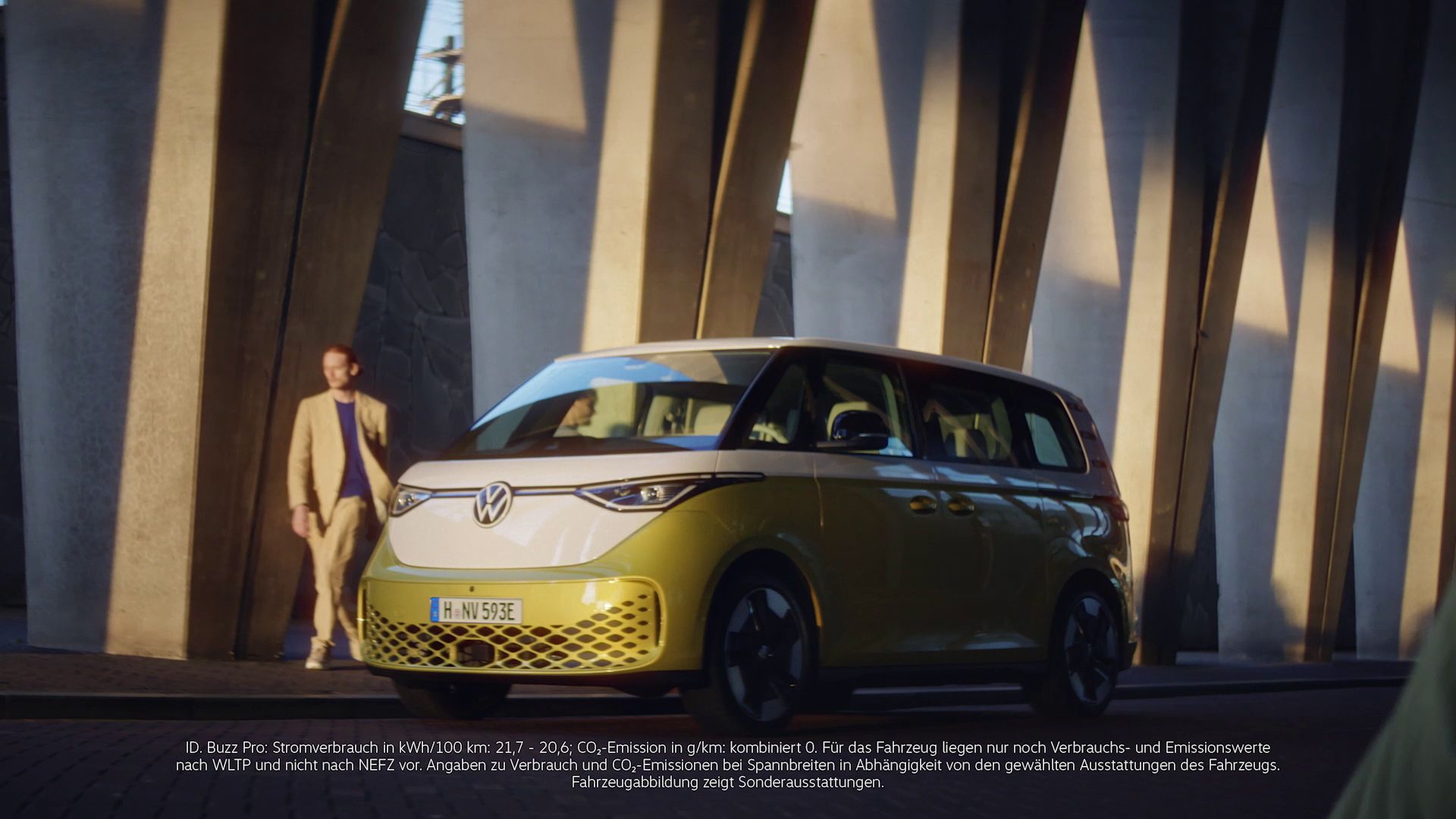 Elektrický ID. Buzz na ulici vedľa moderných architektonických stĺpov. Vozidlo má žltú a bielu farbu. Automobil je zaparkovaný a muž prechádza okolo neho.