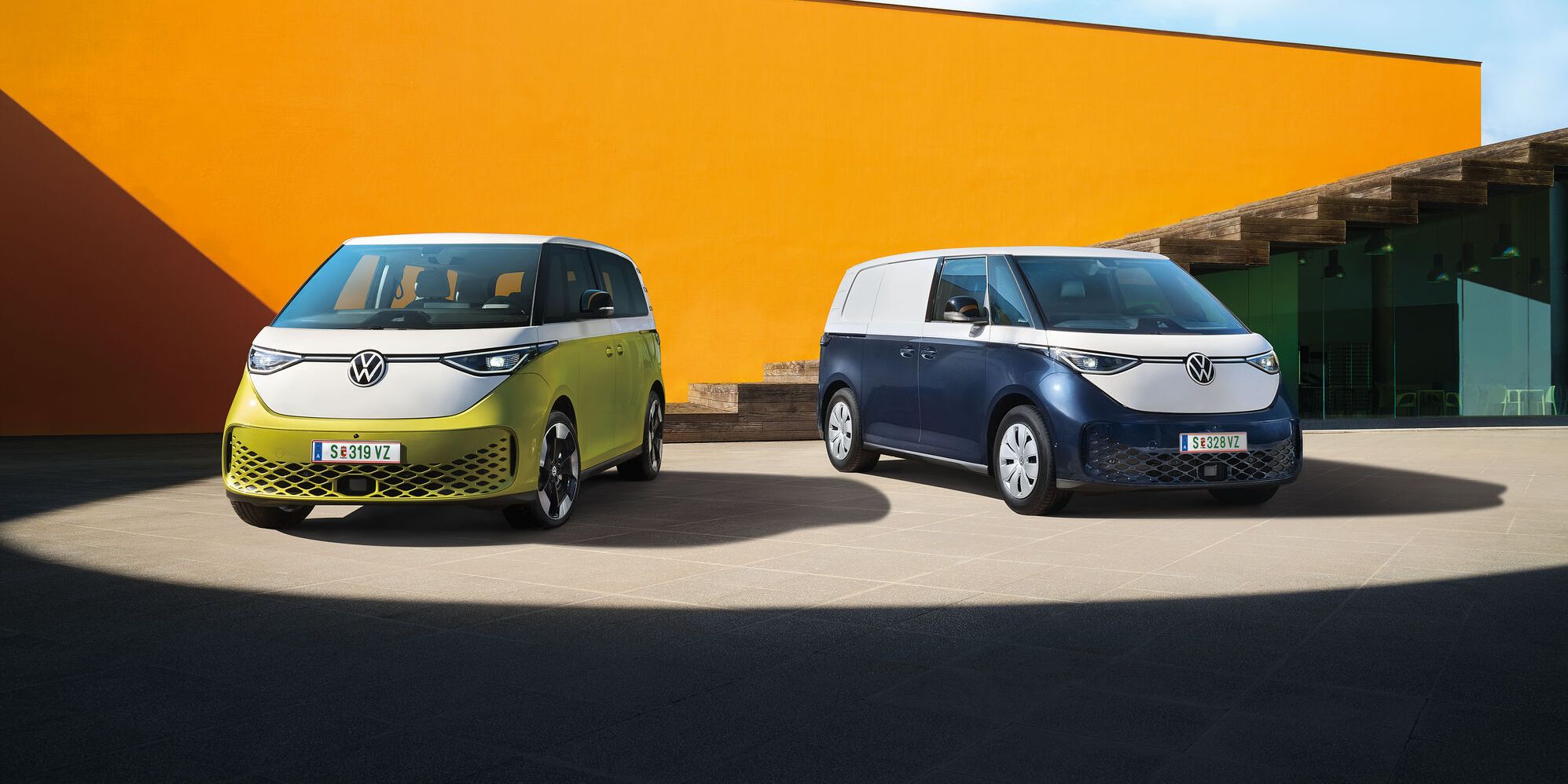 Dva moderné elektrické vozidlá Volkswagen ID.Buzz stoja na betónovom povrchu pred oranžovou stenou modernej budovy. Jedno vozidlo je svetložlté a druhé tmavomodré. Obe majú moderný dizajn s logom VW.