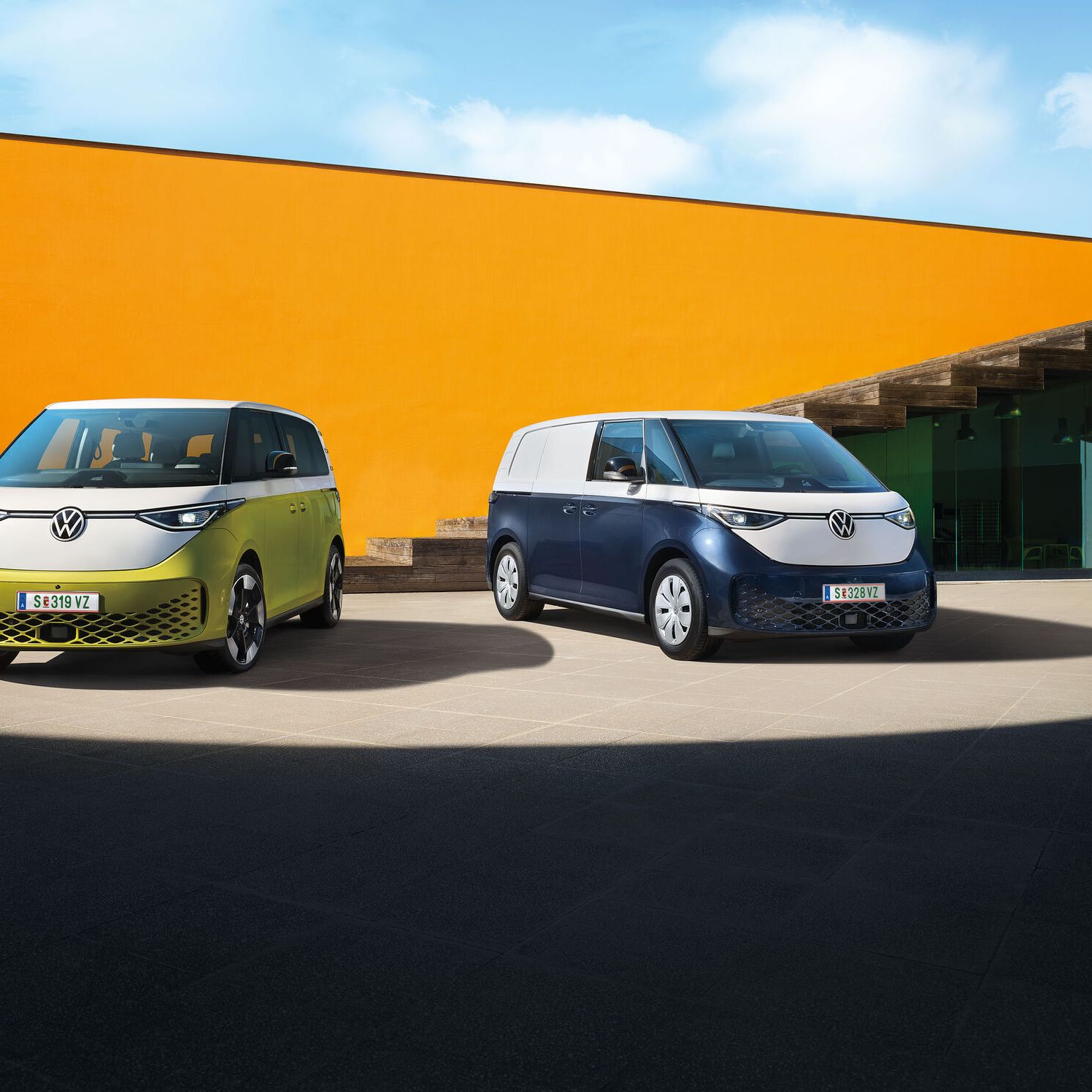 Dva moderné elektrické vozidlá Volkswagen ID.Buzz stoja na betónovom povrchu pred oranžovou stenou modernej budovy. Jedno vozidlo je svetložlté a druhé tmavomodré. Obe majú moderný dizajn s logom VW.