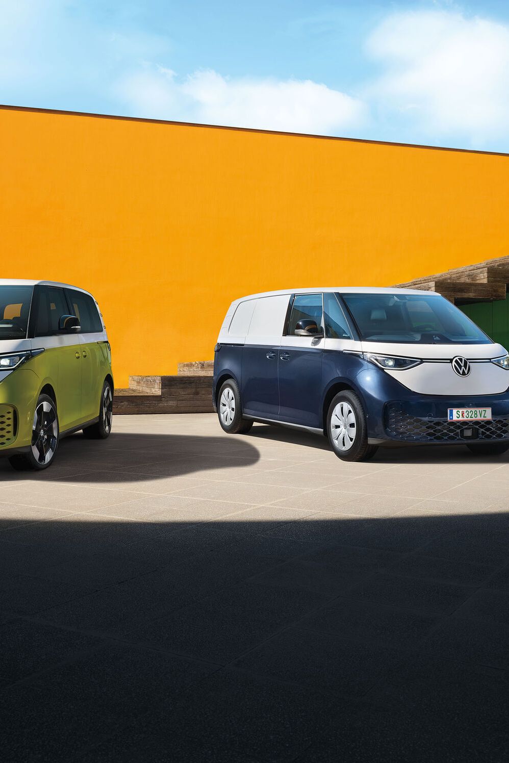 Dva moderné elektrické vozidlá Volkswagen ID.Buzz stoja na betónovom povrchu pred oranžovou stenou modernej budovy. Jedno vozidlo je svetložlté a druhé tmavomodré. Obe majú moderný dizajn s logom VW.