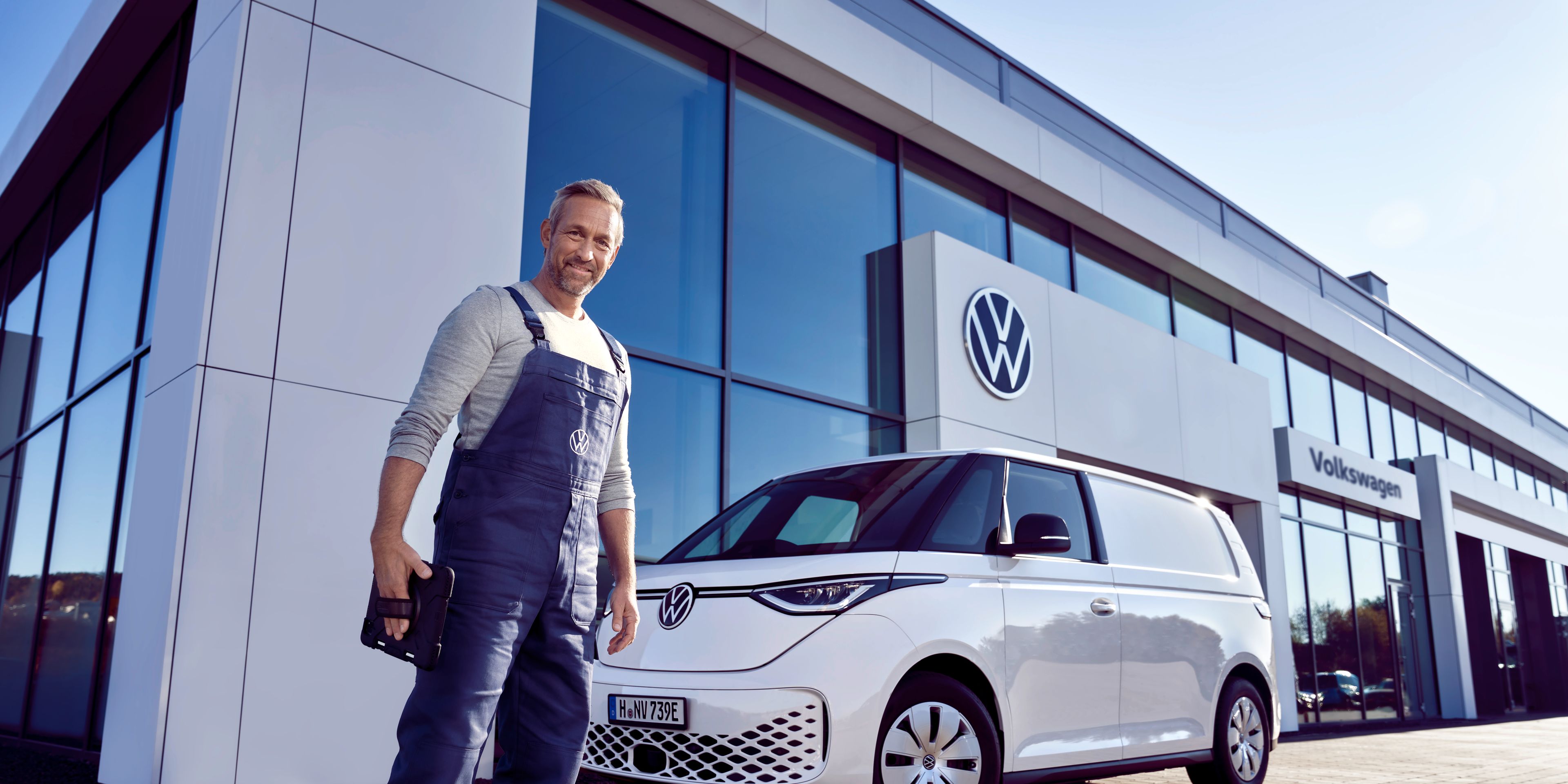 Servisný pracovník v modrých montérkach stojí pred bielym ID. Buzz, ktorí je zaparkovaný pred showroomom Volkswagen. Muž sa pozerá priamo do objektívu.