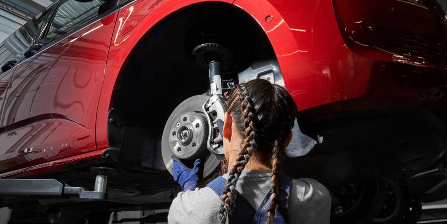 Servisná pracovníčka vykonáva údržbu na prednom kolese červeného auta, ktoré je zdvihnuté. Mechanička kontroluje brzdný kotúč a zavesenie kolies.
