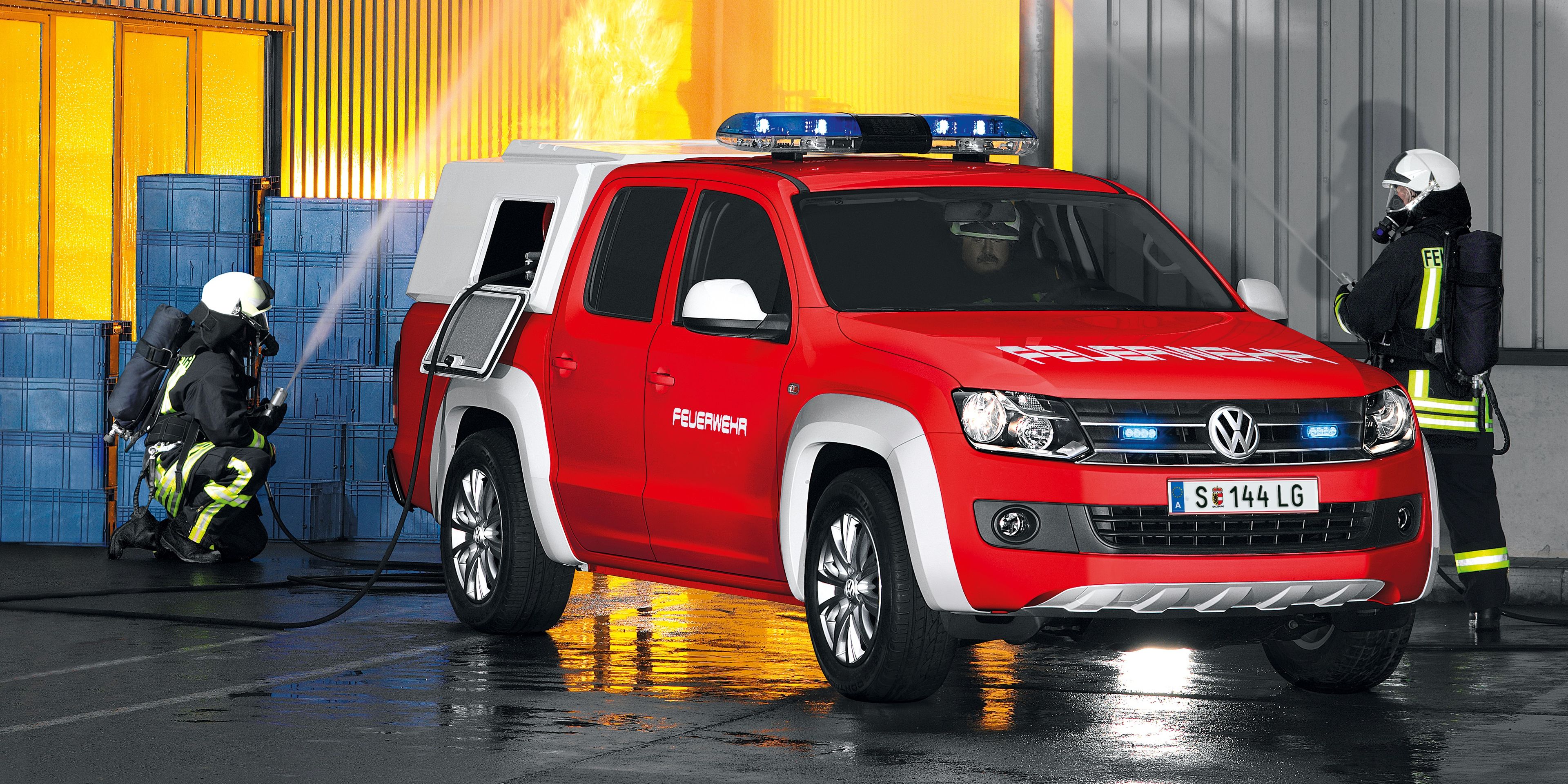 Požiarne auto hasí požiar v priemyselnej budove, z ktorej šľahajú plamene. Dvaja hasiči v ochranných oblekoch manipulujú s hadicami. 