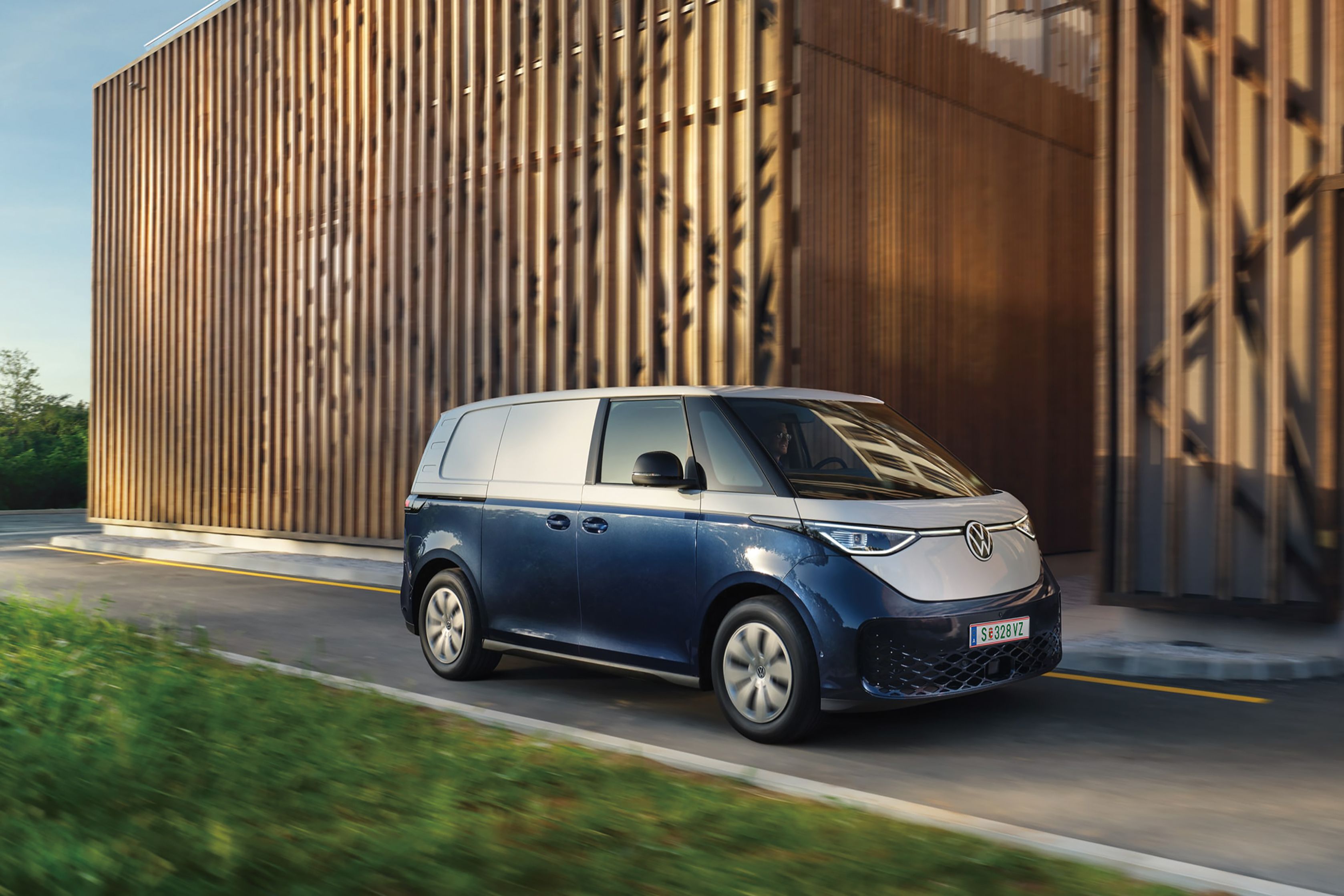 Moderný elektrický modro-biely Volkswagen ID. Buzz Cargo prechádza okolo drevenej budovy za slnečného dňa. Cesta je lemovaná zeleným trávnikom.
