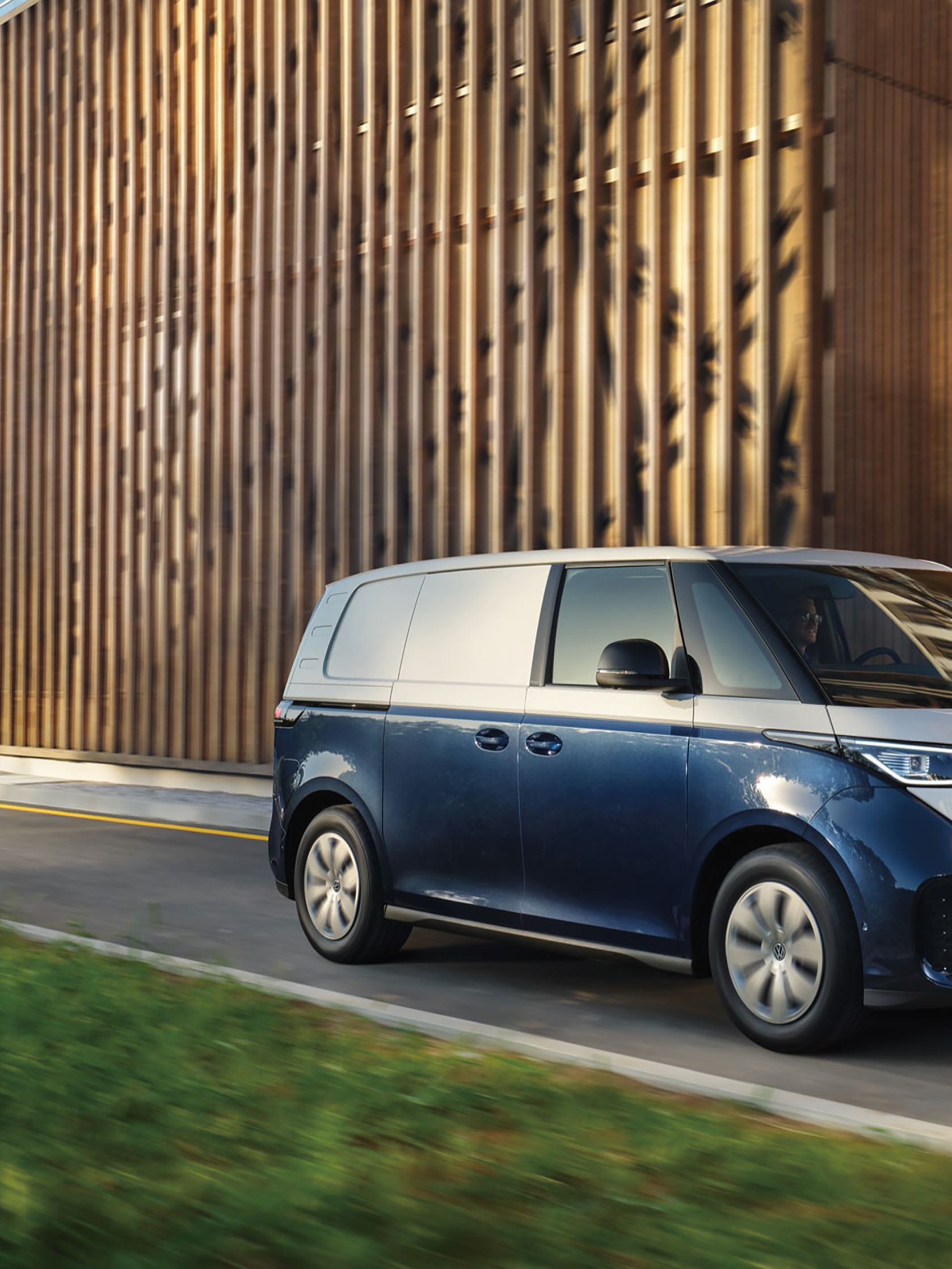 Moderný elektrický modro-biely Volkswagen ID. Buzz Cargo prechádza okolo drevenej budovy za slnečného dňa. Cesta je lemovaná zeleným trávnikom.