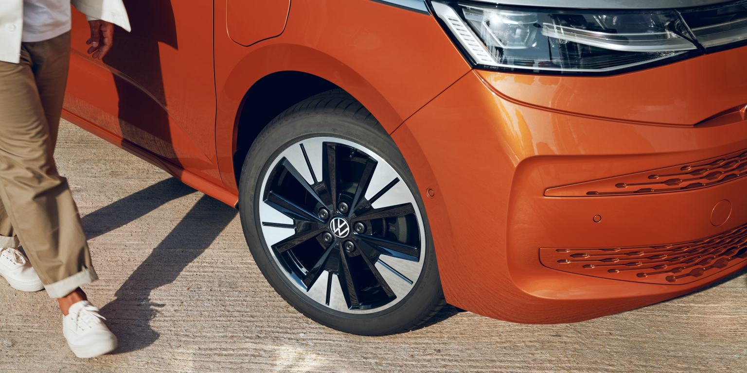 Človek kráča vedľa oranžového auta, z ktorého je vidieť predné koleso. Má na sebe svetlé nohavice a biele tenisky. Na aute je viditeľný znak Volkswagen.