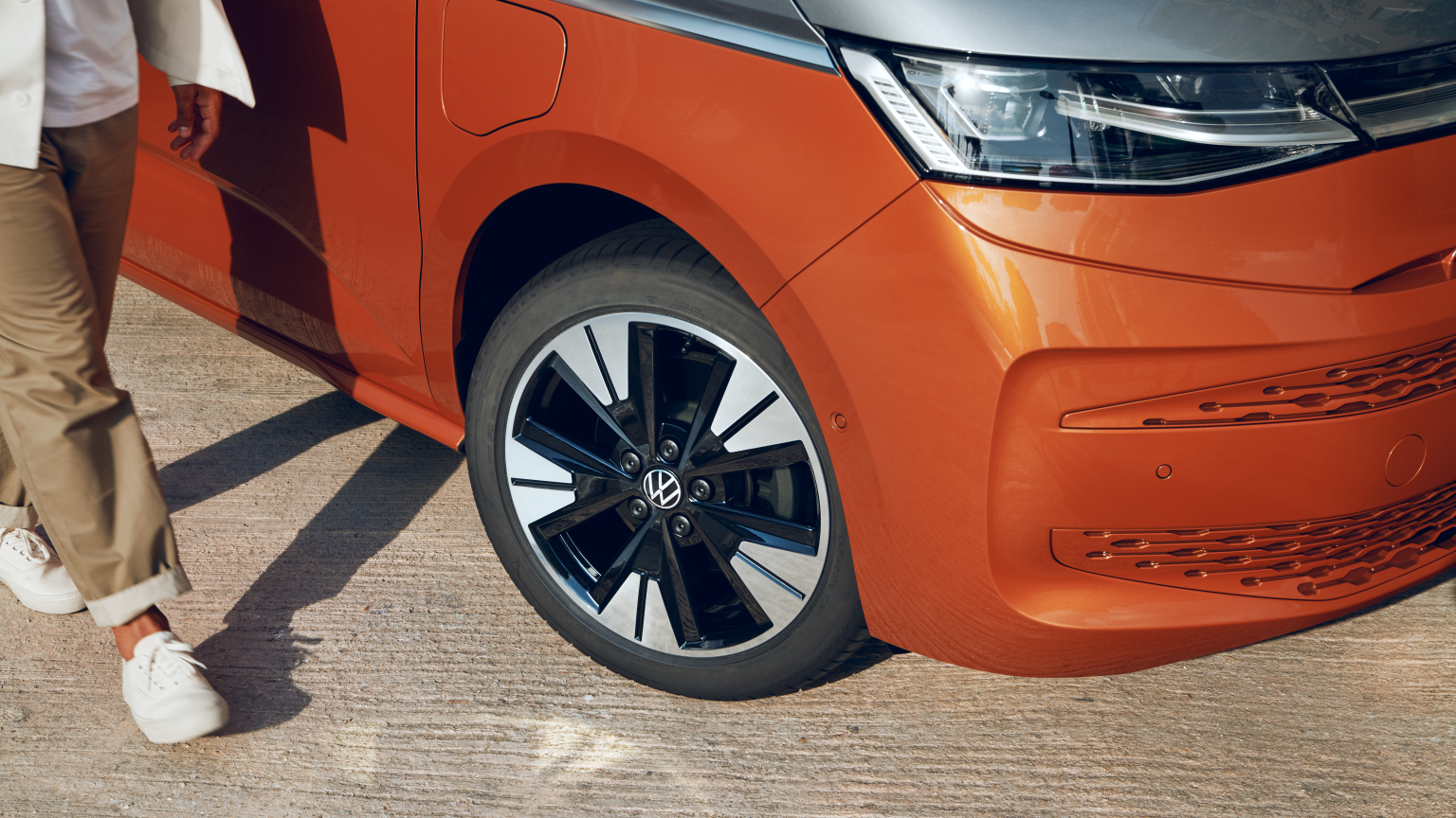 Človek kráča vedľa oranžového auta, z ktorého je vidieť predné koleso. Má na sebe svetlé nohavice a biele tenisky. Na aute je viditeľný znak Volkswagen.