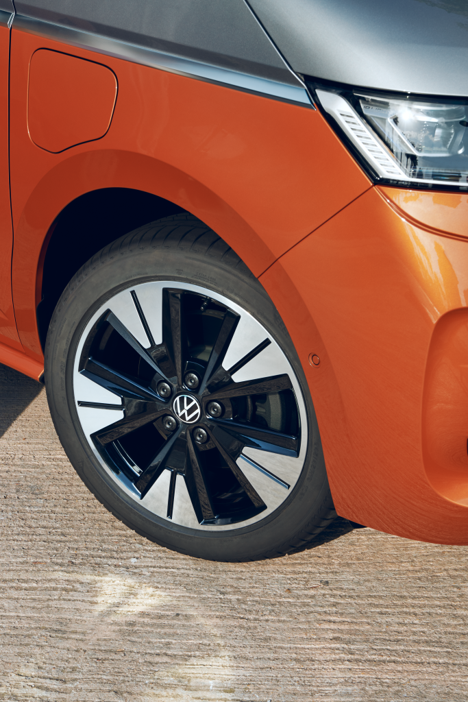 Človek kráča vedľa oranžového auta, z ktorého je vidieť predné koleso. Má na sebe svetlé nohavice a biele tenisky. Na aute je viditeľný znak Volkswagen.