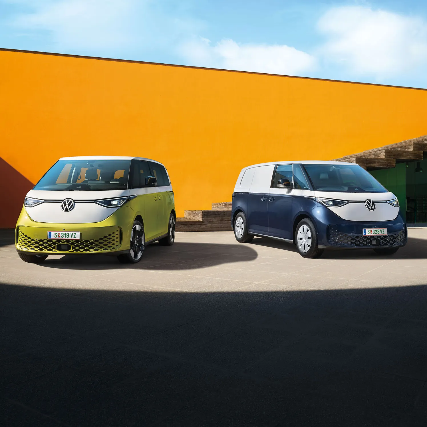Dva moderné elektrické vozidlá Volkswagen ID.Buzz stoja na betónovom povrchu pred oranžovou stenou modernej budovy. Jedno vozidlo je svetložlté a druhé tmavomodré. Obe majú moderný dizajn s logom VW.