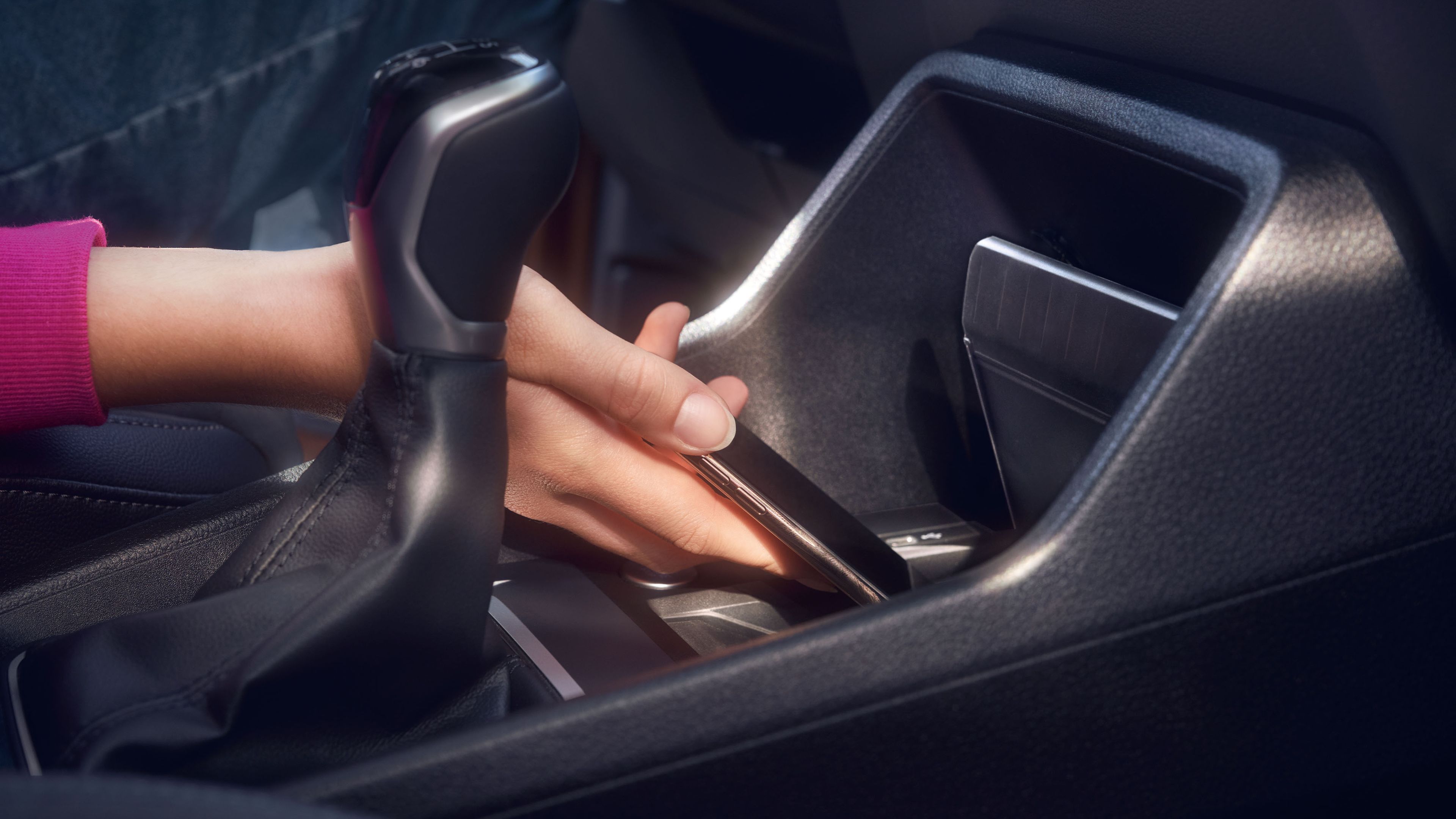 Ruka drži mobilný telefón v aute a ukladá ho do odkladacej priehradky pre bezdrôtové nabíjanie. Vo vnútri sú viditeľné moderné ovládacie prvky automobilu.