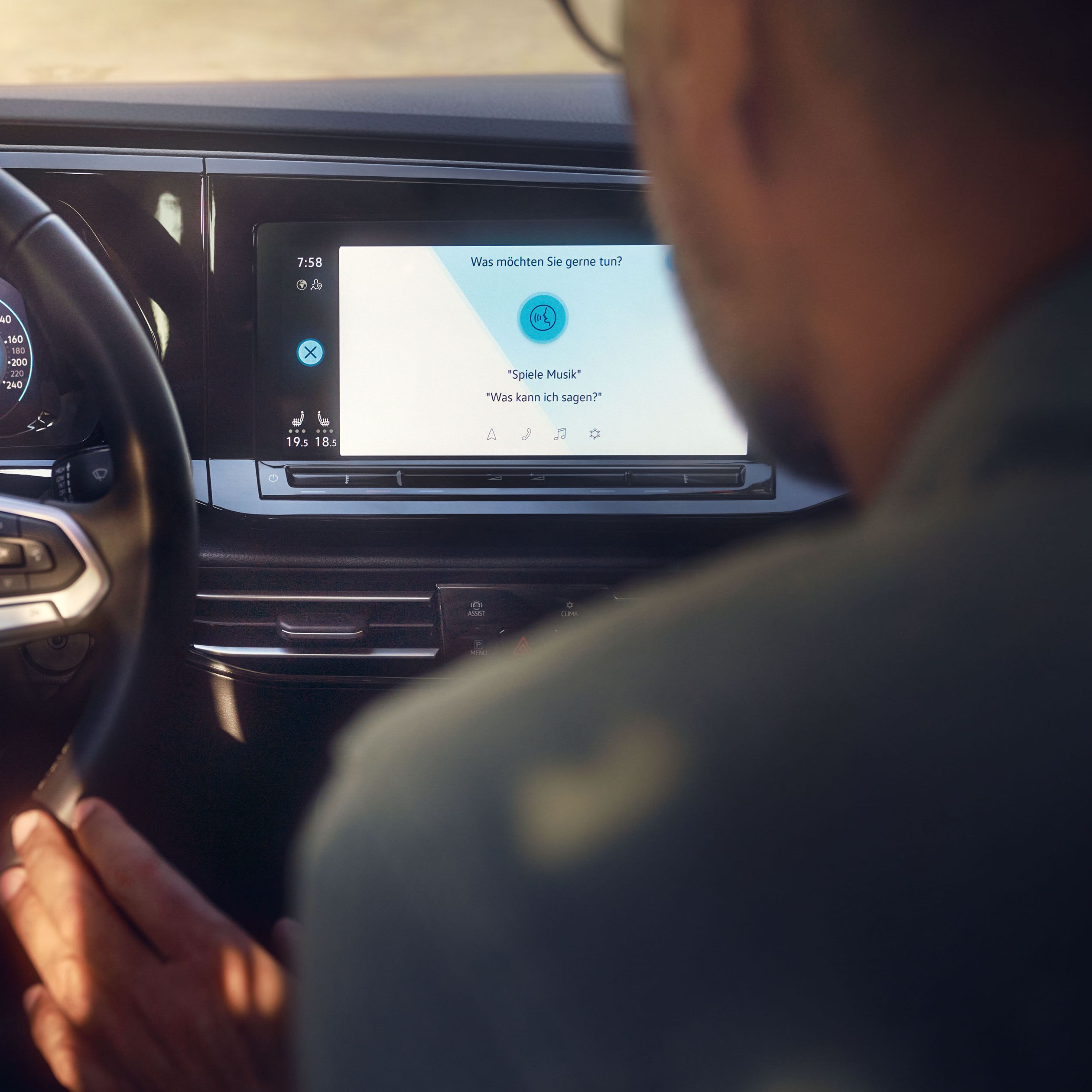 Muž sedí na mieste spolujazdca a dotýka sa volantu, ovláda systém infotainment prostredníctvom dotykového displeja. Na obrazovke vidieť aktivované hlasové ovládanie. Interiér vozidla pôsobí moderným a technologicky pokročilým dojmom.