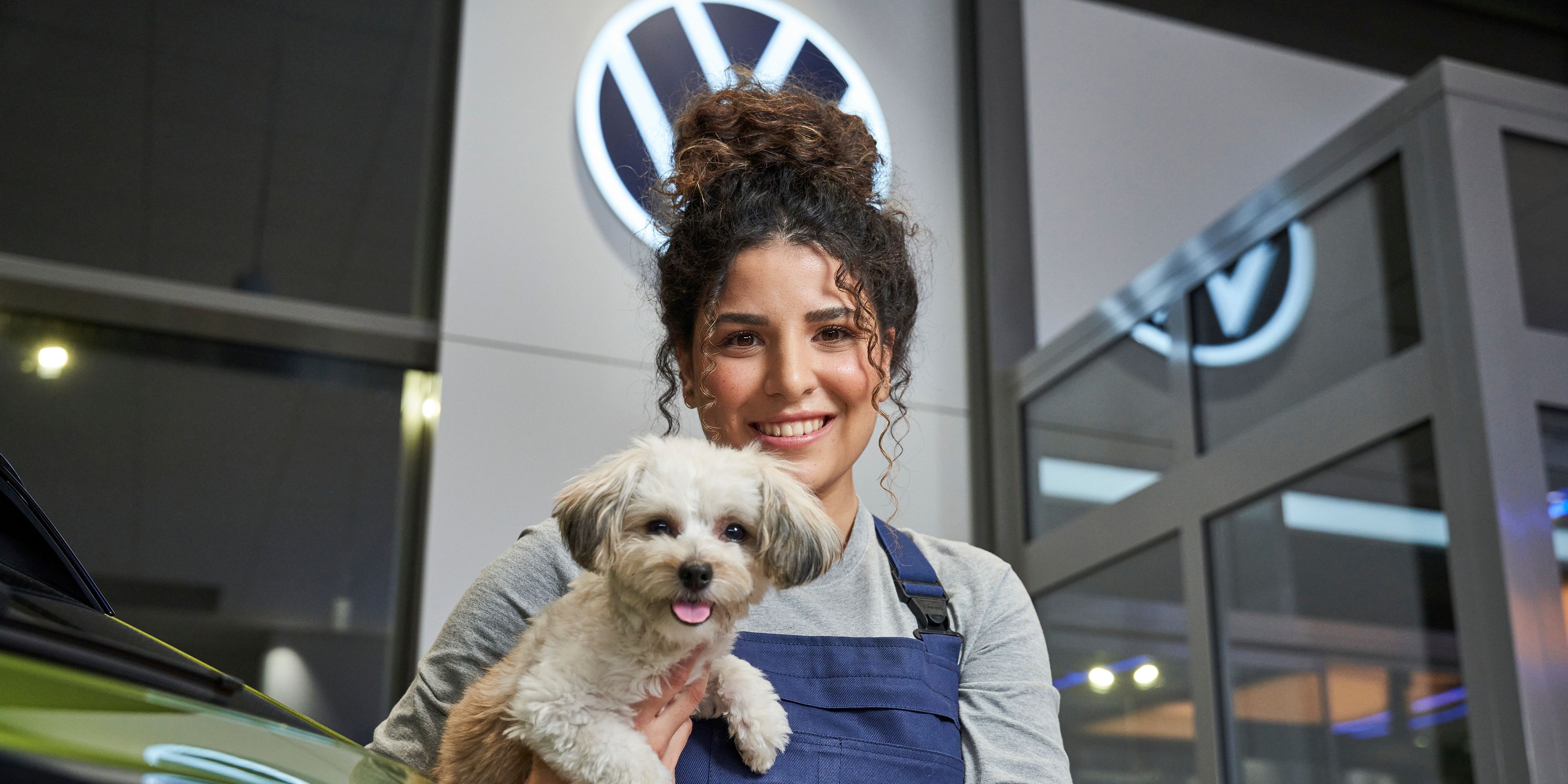 Servisná pracovníčka v modrom pracovnom oblečení drží malého psa na rukách. V pozadí je viditeľné logo Volkswagen. Scéna sa odohráva v autosalóne.