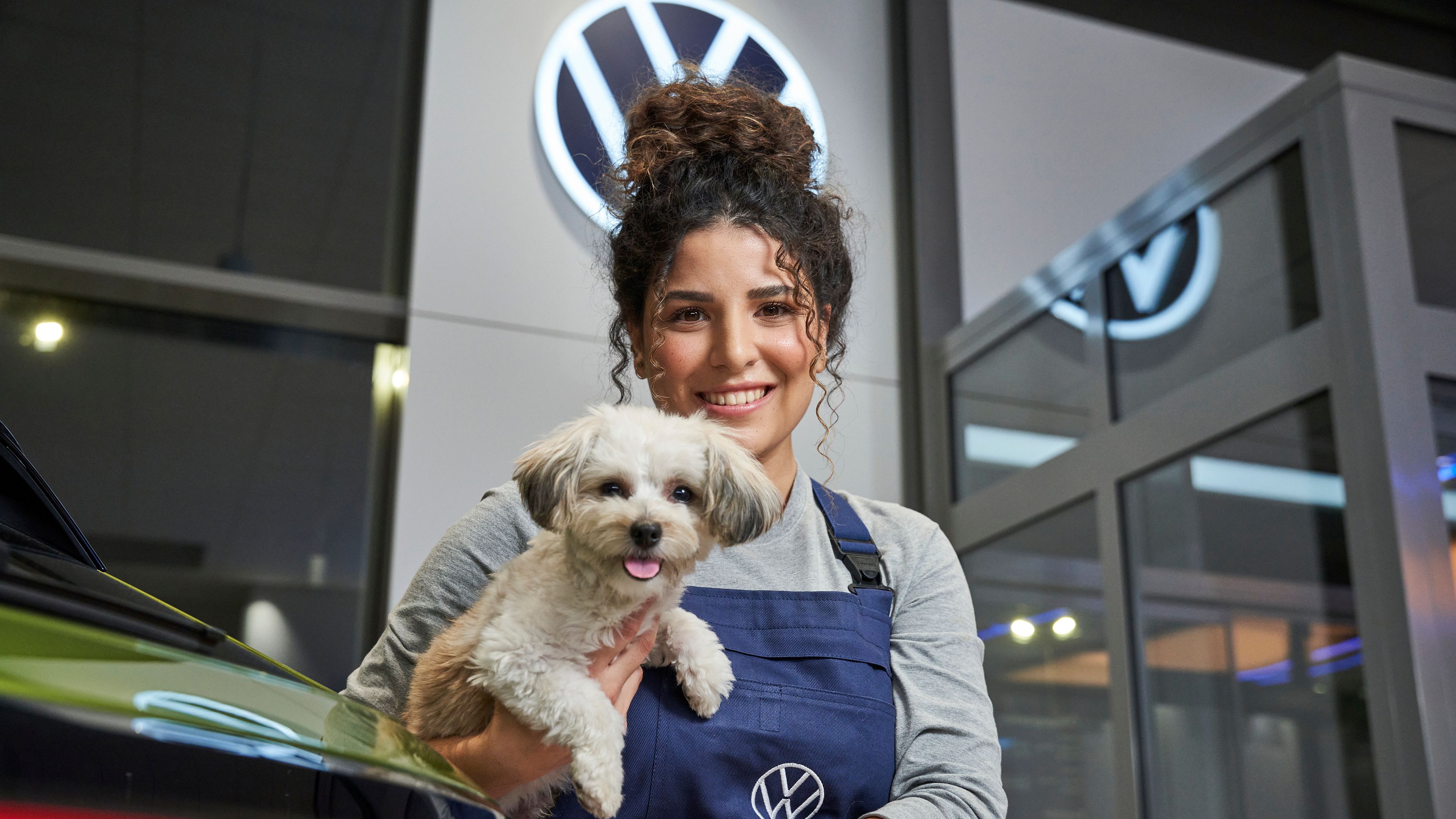 Servisná pracovníčka v modrom pracovnom oblečení drží malého psa na rukách. V pozadí je viditeľné logo Volkswagen. Scéna sa odohráva v autosalóne.
