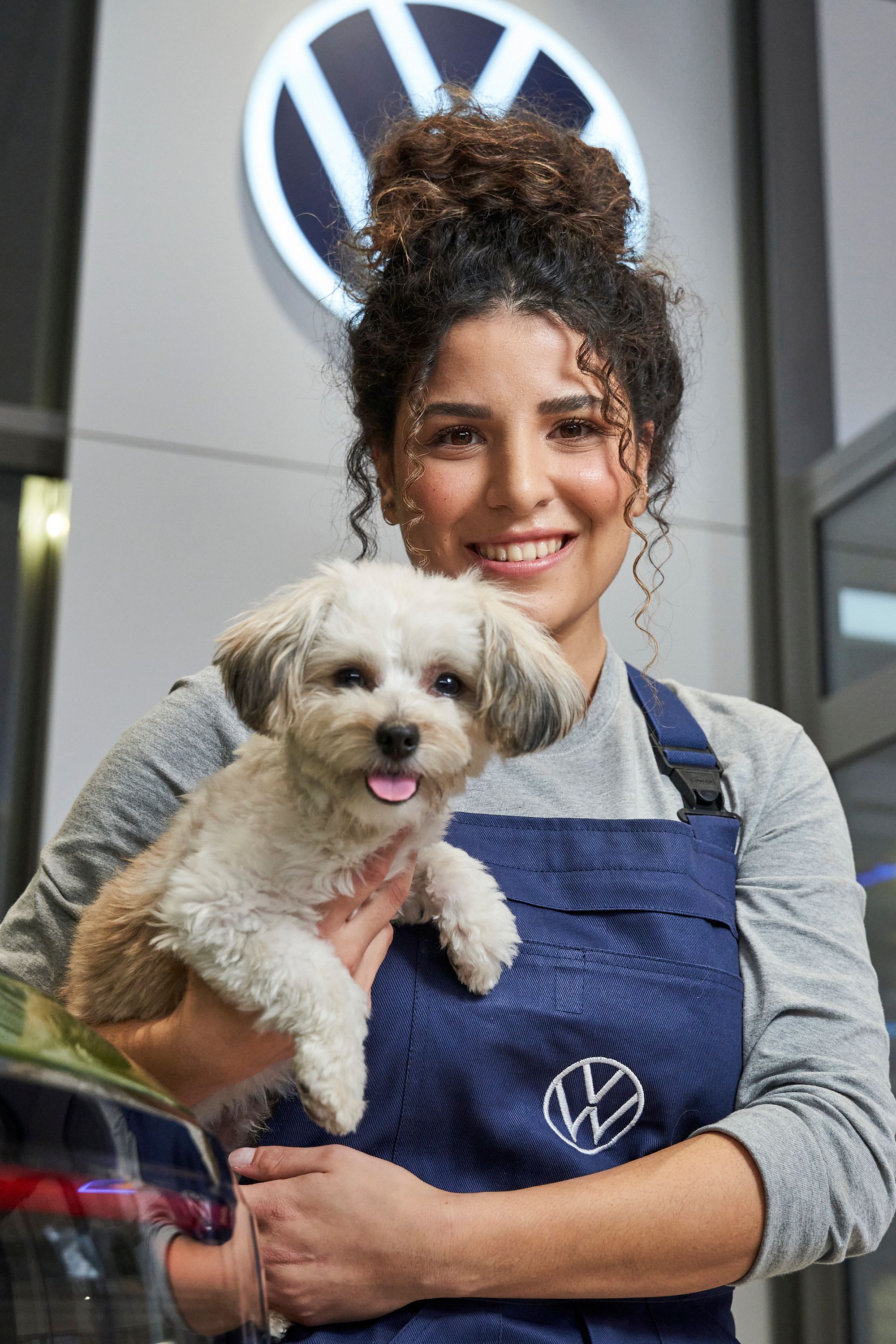 Servisná pracovníčka v modrom pracovnom oblečení drží malého psa na rukách. V pozadí je viditeľné logo Volkswagen. Scéna sa odohráva v autosalóne.
