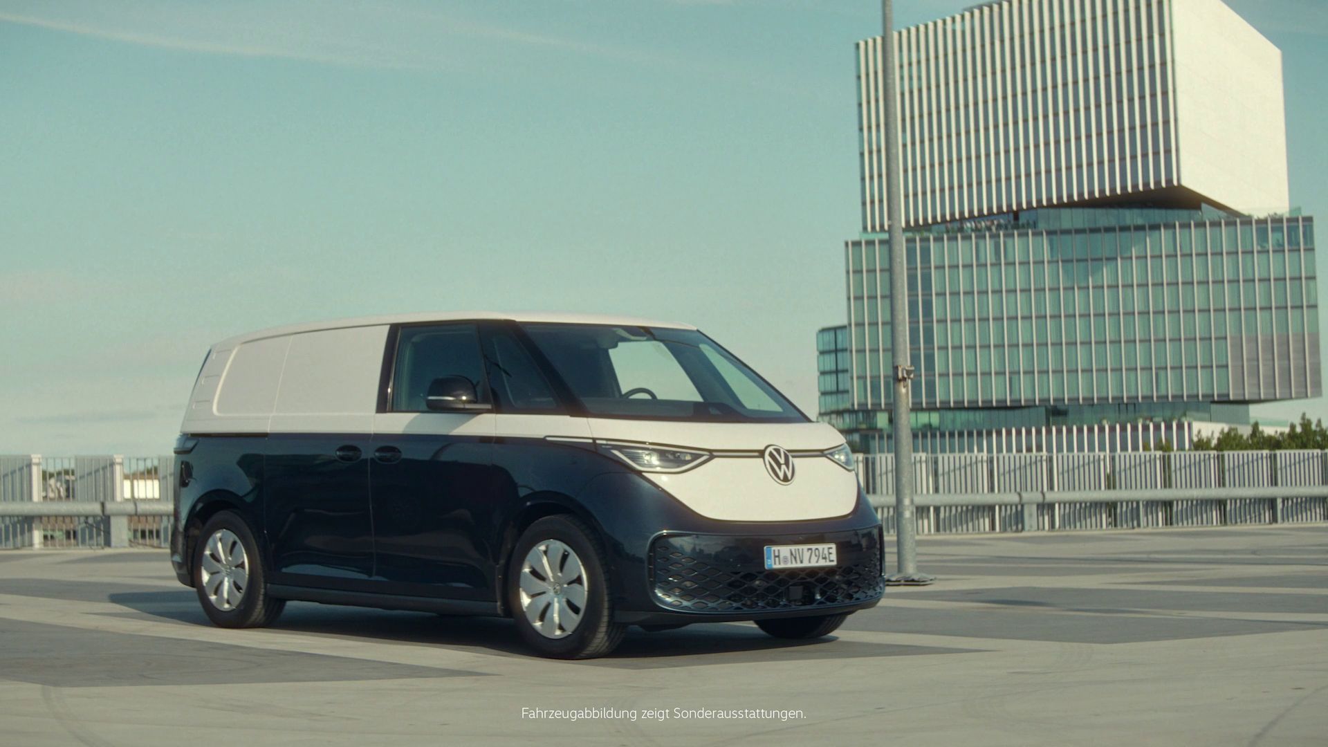 ID. Buzz Cargo zaparkovaný na parkovisku. V pozadí je vysoká moderná budova. Obloha je modrá a jemnými mrakmi.