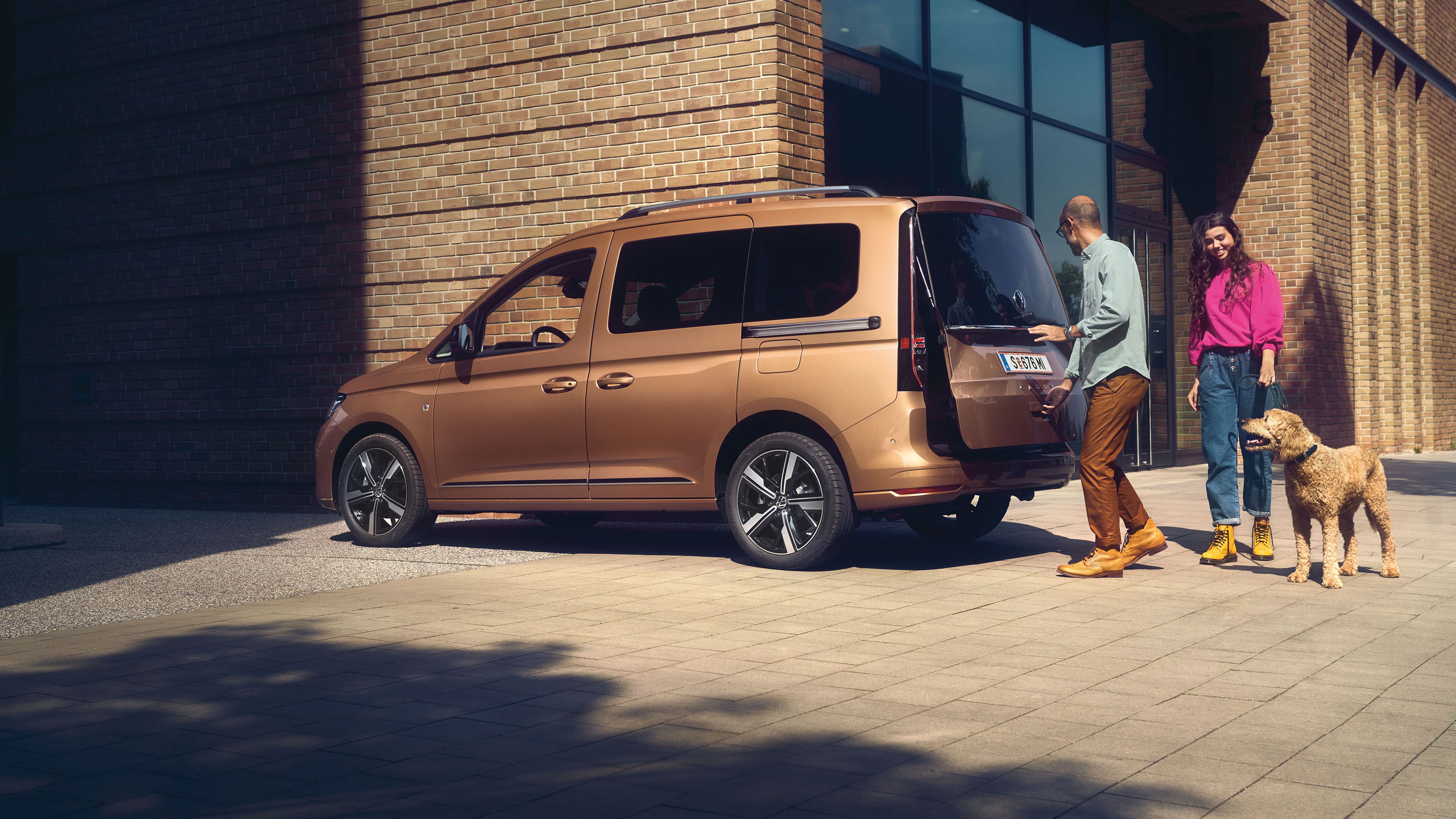 Muž a žena so psom stoja pri VW Caddy. Muž otvára dvere batožinového priestoru hnedého VW Caddy zaparkovaného pred modernou, tehlovou budovou.