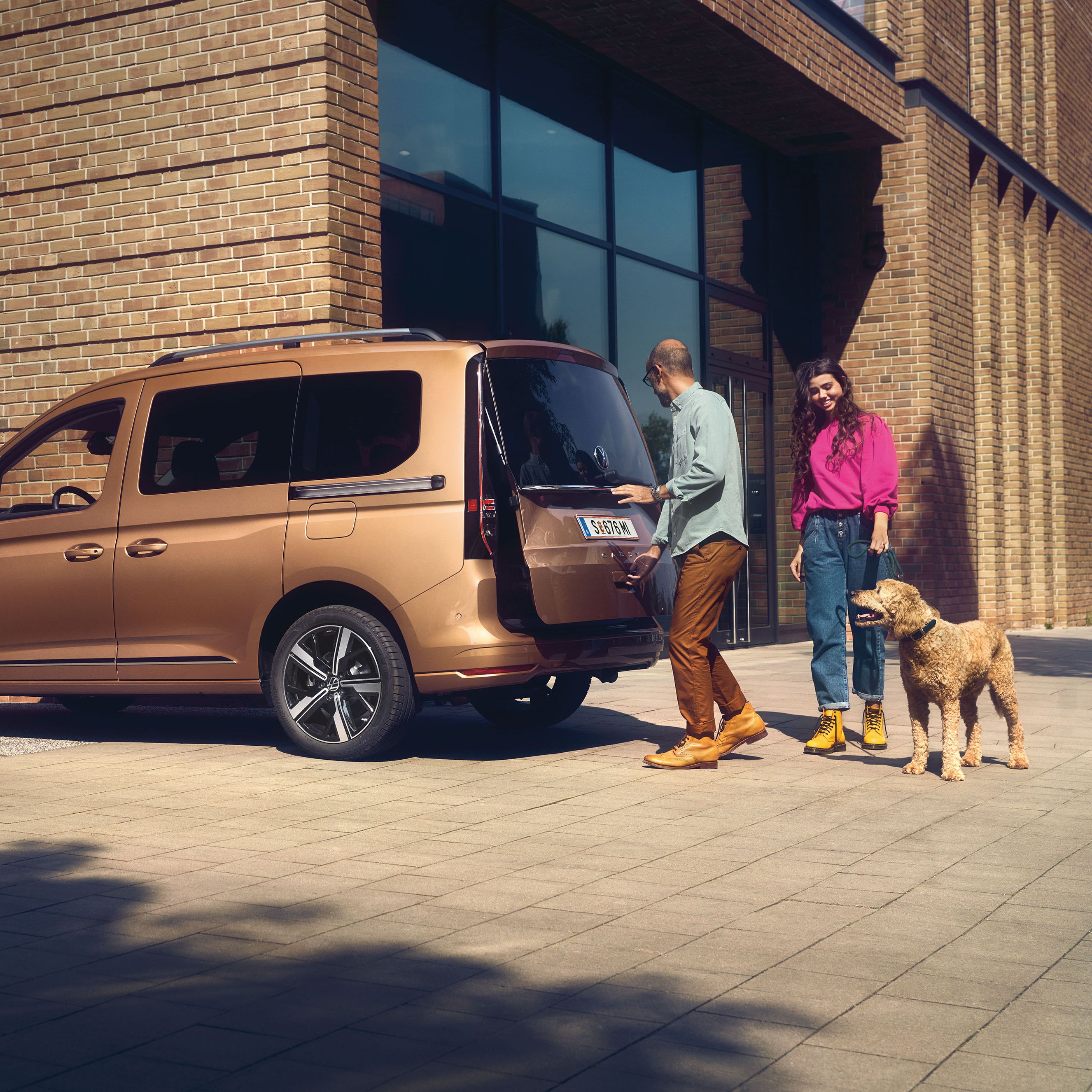 Muž a žena so psom stoja pri VW Caddy. Muž otvára dvere batožinového priestoru hnedého VW Caddy zaparkovaného pred modernou, tehlovou budovou.