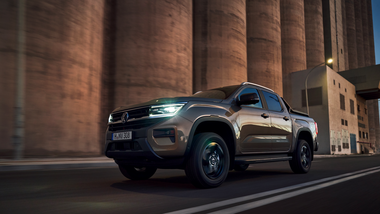 Tmavohnedý pickup Volkswagen jazdí po ceste pred veľkými priemyselnými budovami. Svetlá automobilu sú zapnuté a okolie je mierne osvetlené.