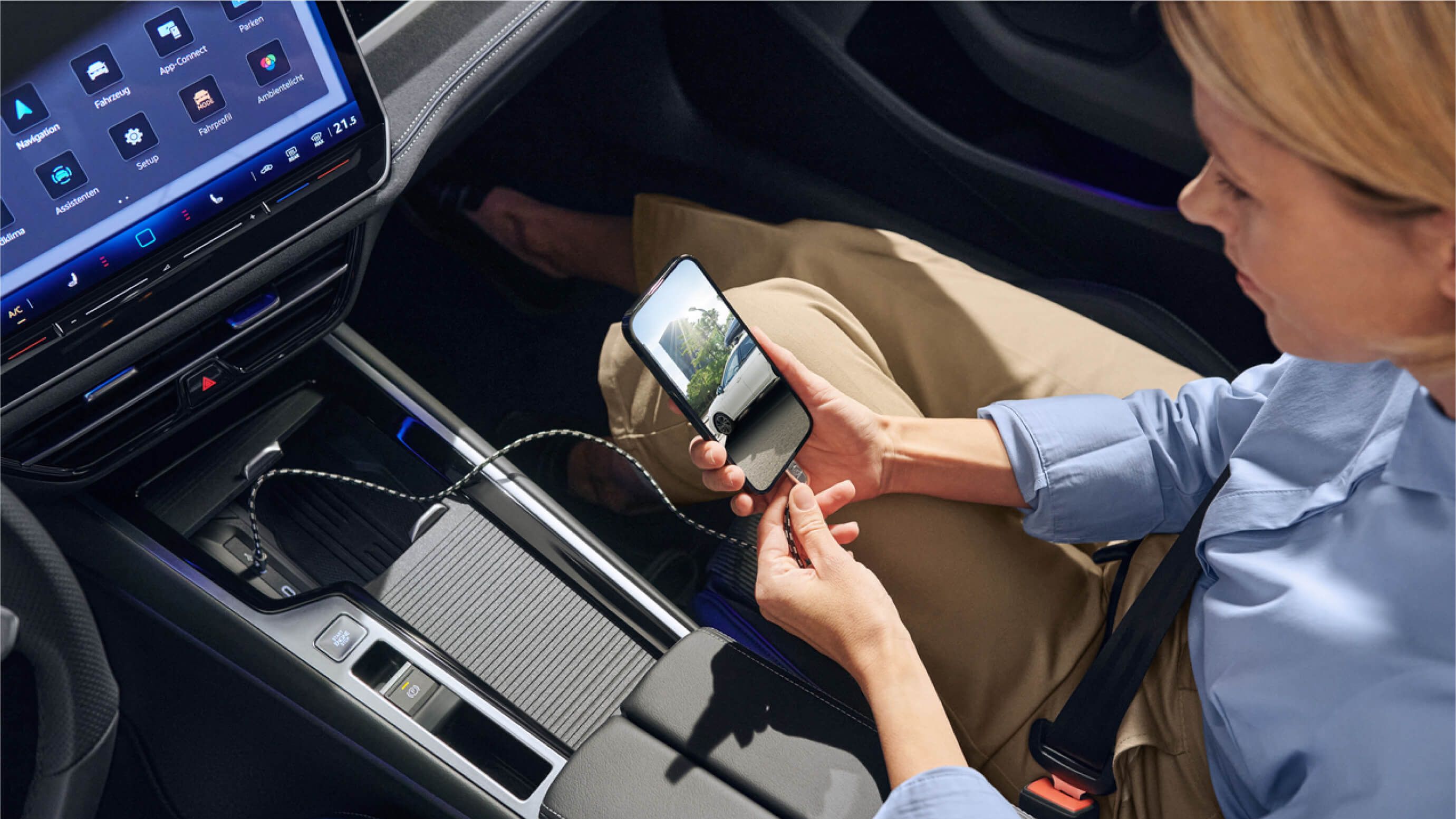 Žena sedí v aute a drží mobilný telefón pripojený USB káblom k vozidlu, aby sa nabíjal. Na displeji mobilu je vidieť cestu, ktorou prechádza auto. Na palubnej doske sú tiež viditeľné dotykové ovládacie panely a displej systému infotainment.