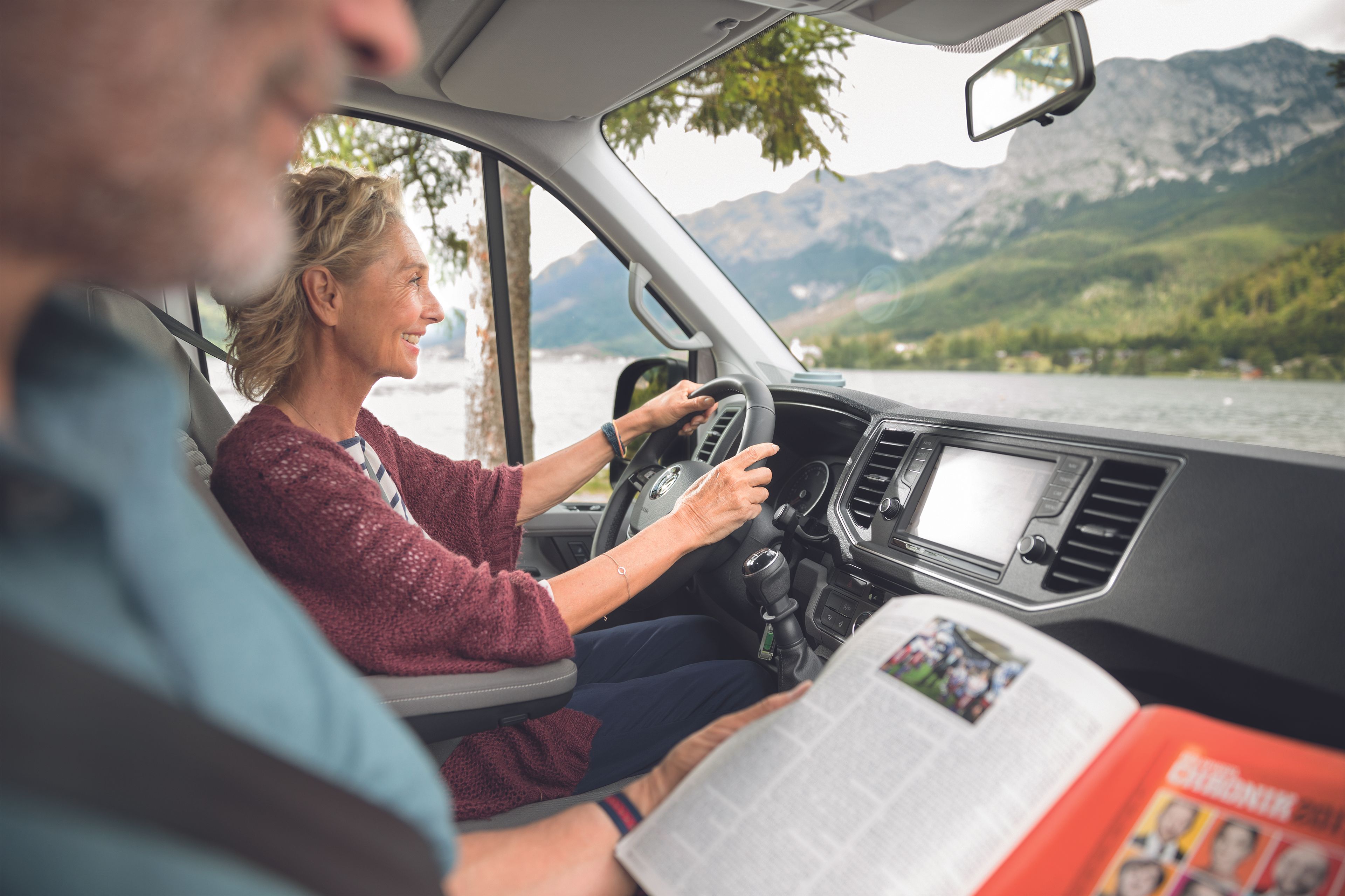 Dvojica sedí v Grand California. Žena riadi vozidlo a usmieva sa pritom. Muž sedí vedľa nej a číta si časopis.