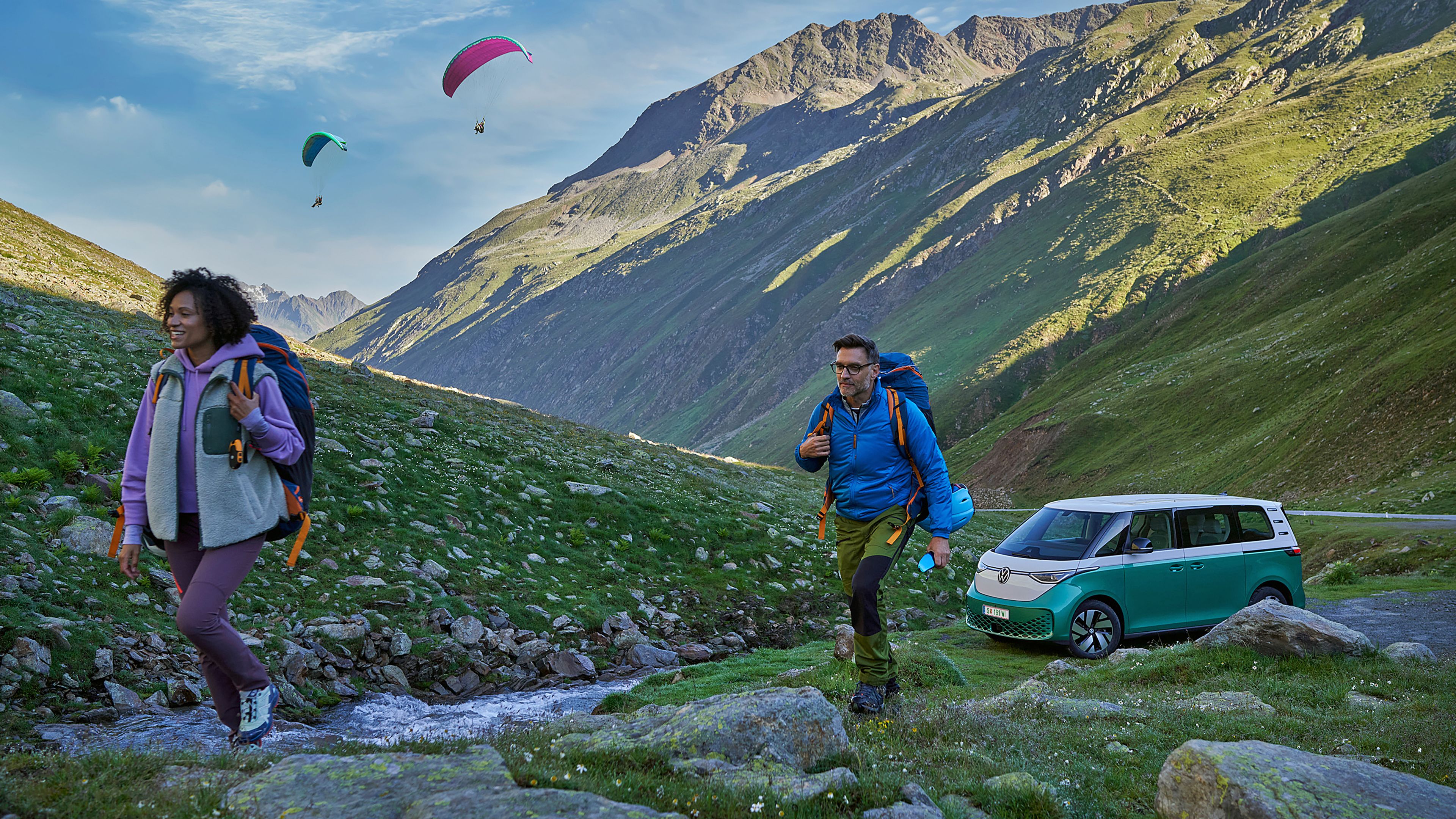 Dvaja turisti so zabalenými batohmi kráčajú po horskom údolí plnom zelene blízko potoka. Za nimi vidieť dvoch paraglidistov vo vzduchu a zelený ID. Buzz je zaparkovaný vedľa cesty pri potôčiku. V pozadí vidieť hory a skalnatý terén.