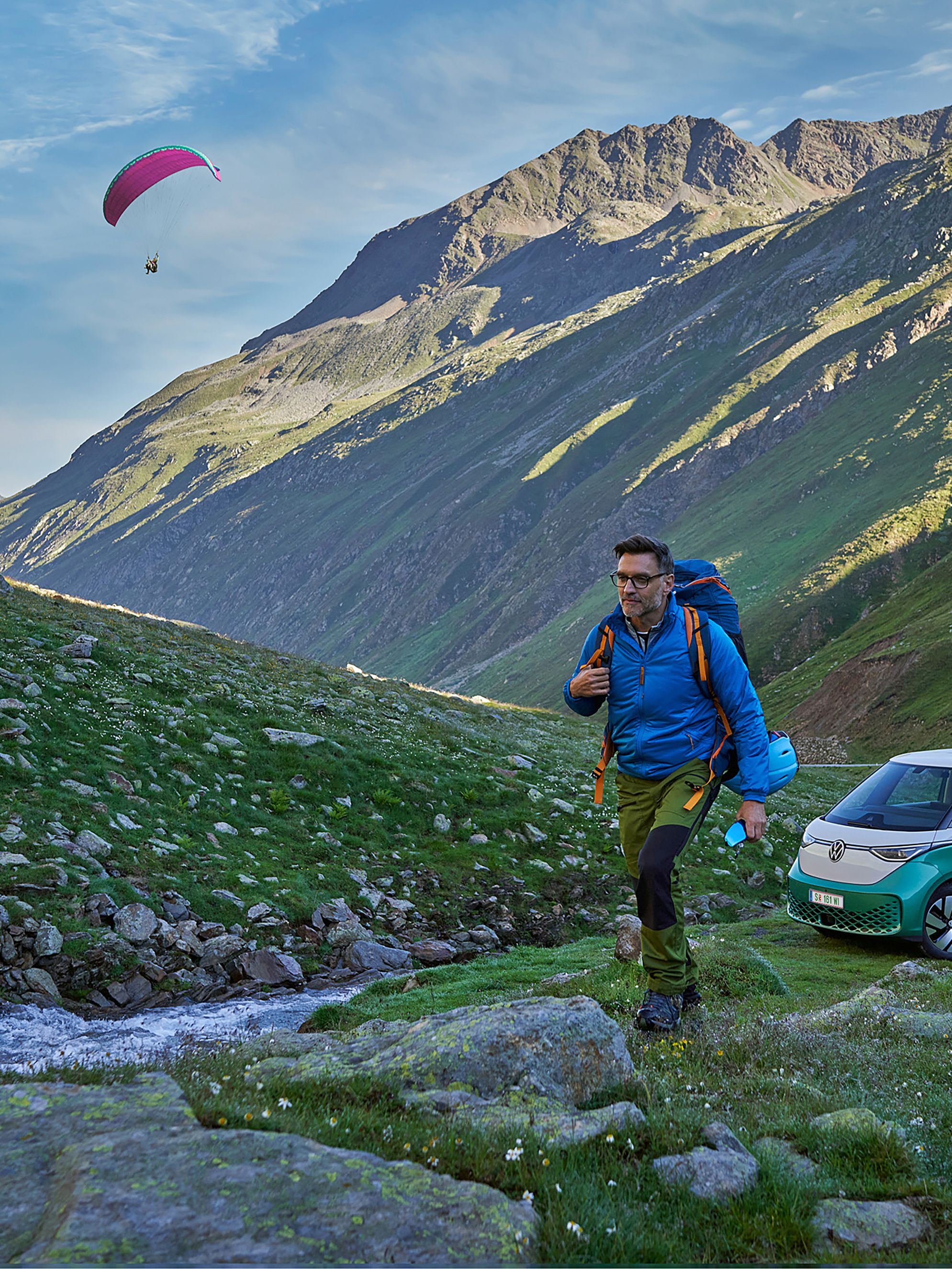 Dvaja turisti so zabalenými batohmi kráčajú po horskom údolí plnom zelene blízko potoka. Za nimi vidieť dvoch paraglidistov vo vzduchu a zelený ID. Buzz je zaparkovaný vedľa cesty pri potôčiku. V pozadí vidieť hory a skalnatý terén.