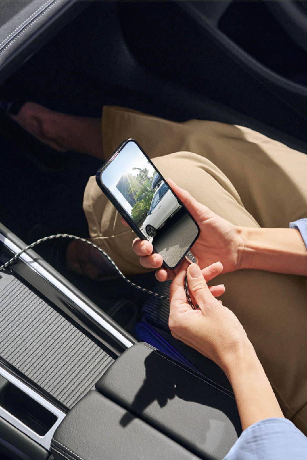 Žena sedí v aute a drží mobilný telefón pripojený USB káblom k vozidlu, aby sa nabíjal. Na displeji mobilu je vidieť cestu, ktorou prechádza auto. Na palubnej doske sú tiež viditeľné dotykové ovládacie panely a displej systému infotainment.