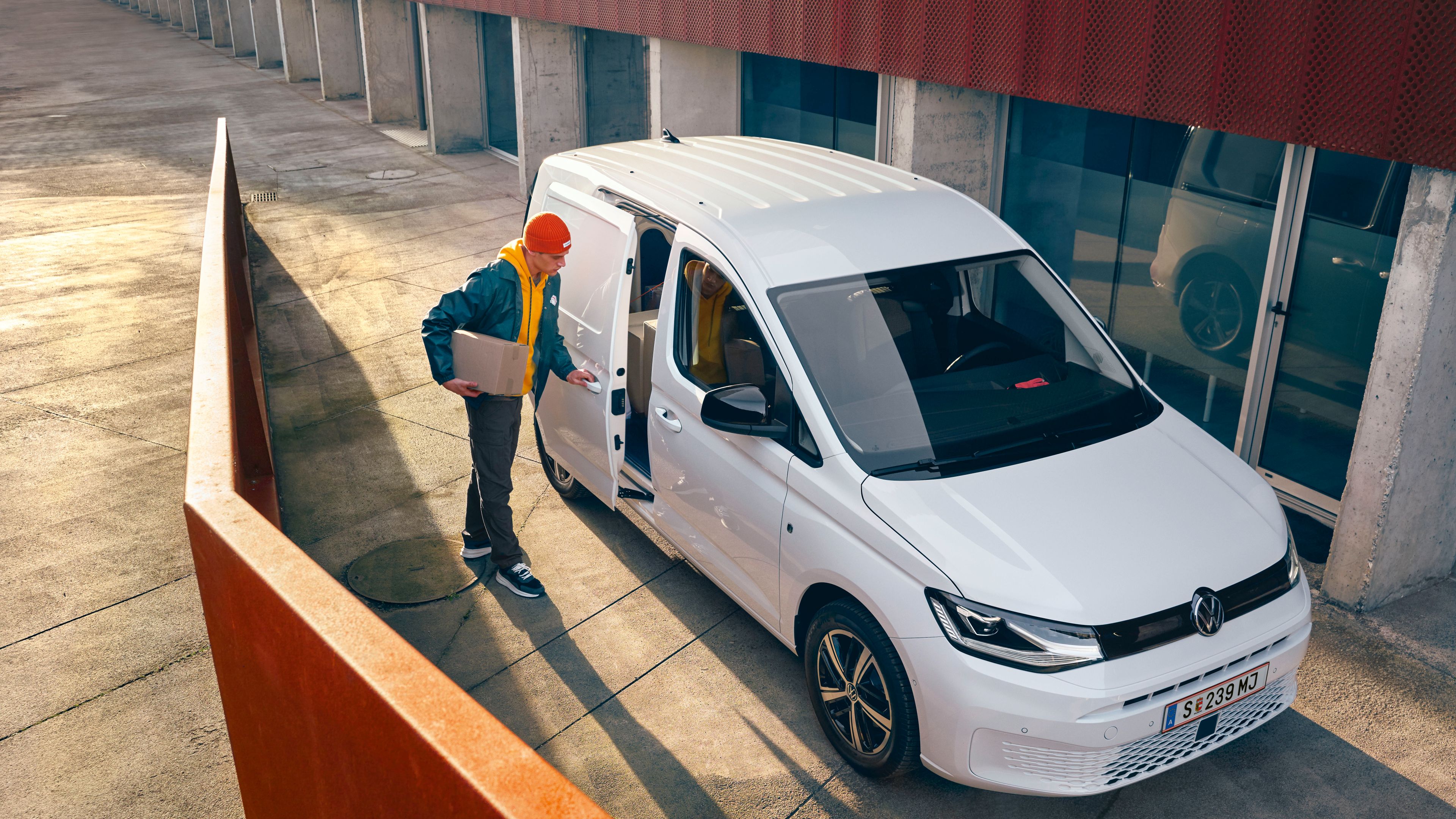 Osoba s krabicou v ruke zatvára bočné dvere bielej dodávky VW Caddy Cargo zaparkovanej pri modernej budove s veľkými oknami 