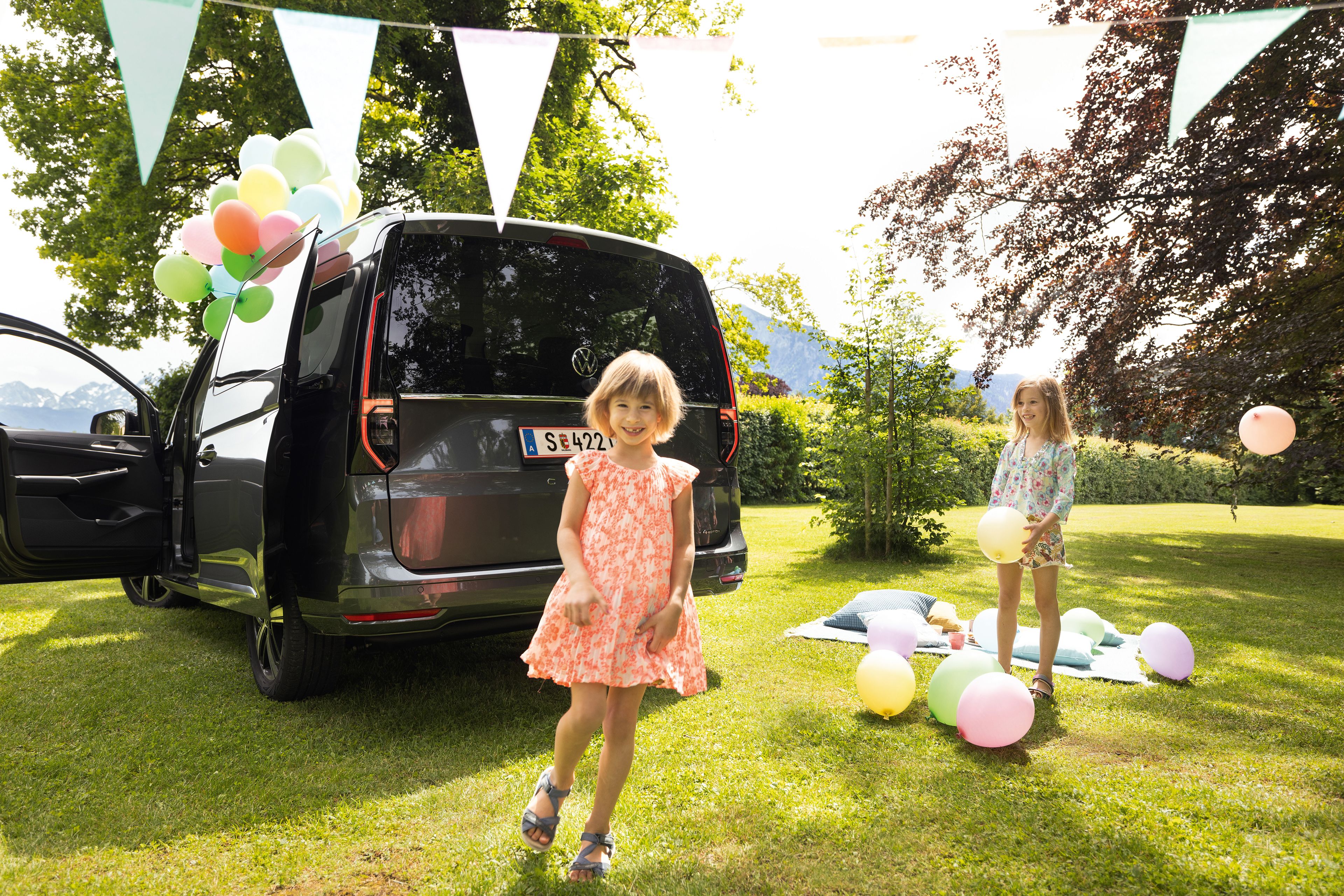 Dve malé dievčatá na narodeninovej oslave ako piknik v parku. Auto je zaparkované bokom a má otvorené dvere, z vozidla trčia farebné balóny, ktoré sú aj na lúke a na deke.  Slnečný deň dotvára radostnú atmosféru.