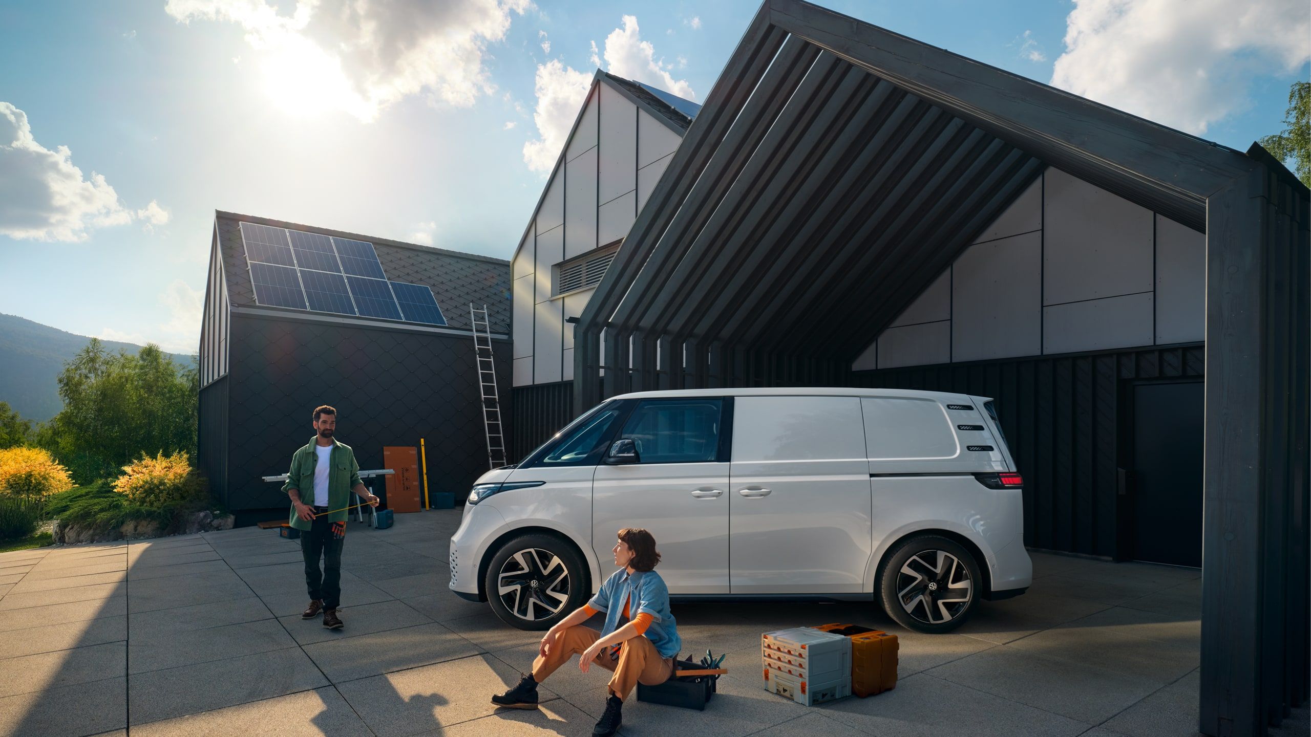 Biely ID. Buzz Cargo zaparkovaný pred moderným domom. Muž prichádza okolo dodávky a približuje sa žene sediacej na krabici s nástrojmi vedľa vozidla. V pozadí je moderná budova so solárnymi panelmi na streche.