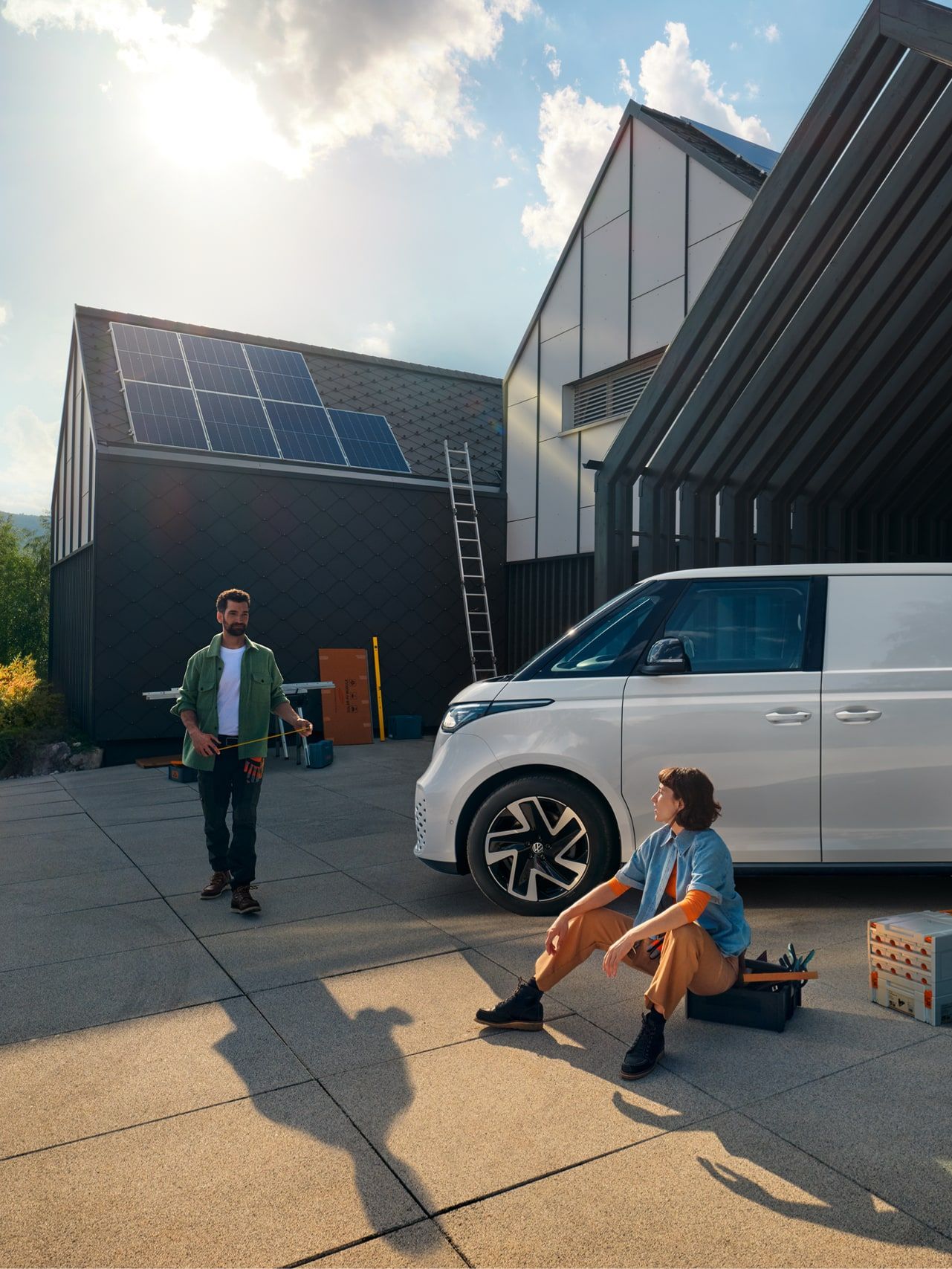 Biely ID. Buzz Cargo zaparkovaný pred moderným domom. Muž prichádza okolo dodávky a približuje sa žene sediacej na krabici s nástrojmi vedľa vozidla. V pozadí je moderná budova so solárnymi panelmi na streche.